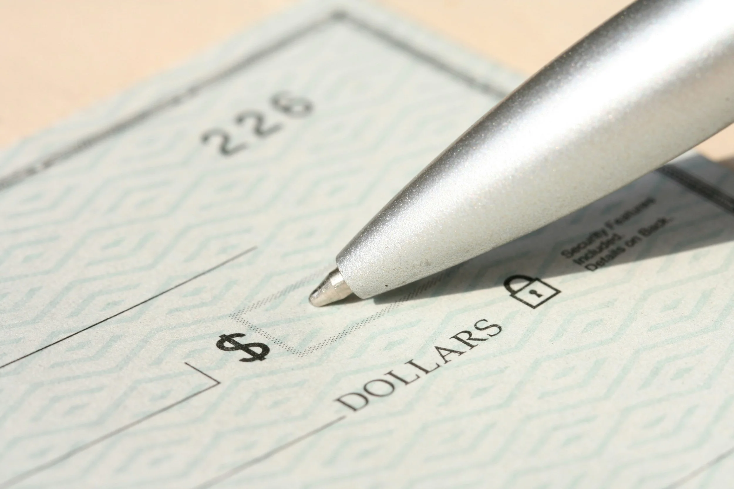 A close-up image of a blank check showing the dollar sign box and part of the “DOLLARS” line, with a metallic ballpoint pen rests near the writing area.