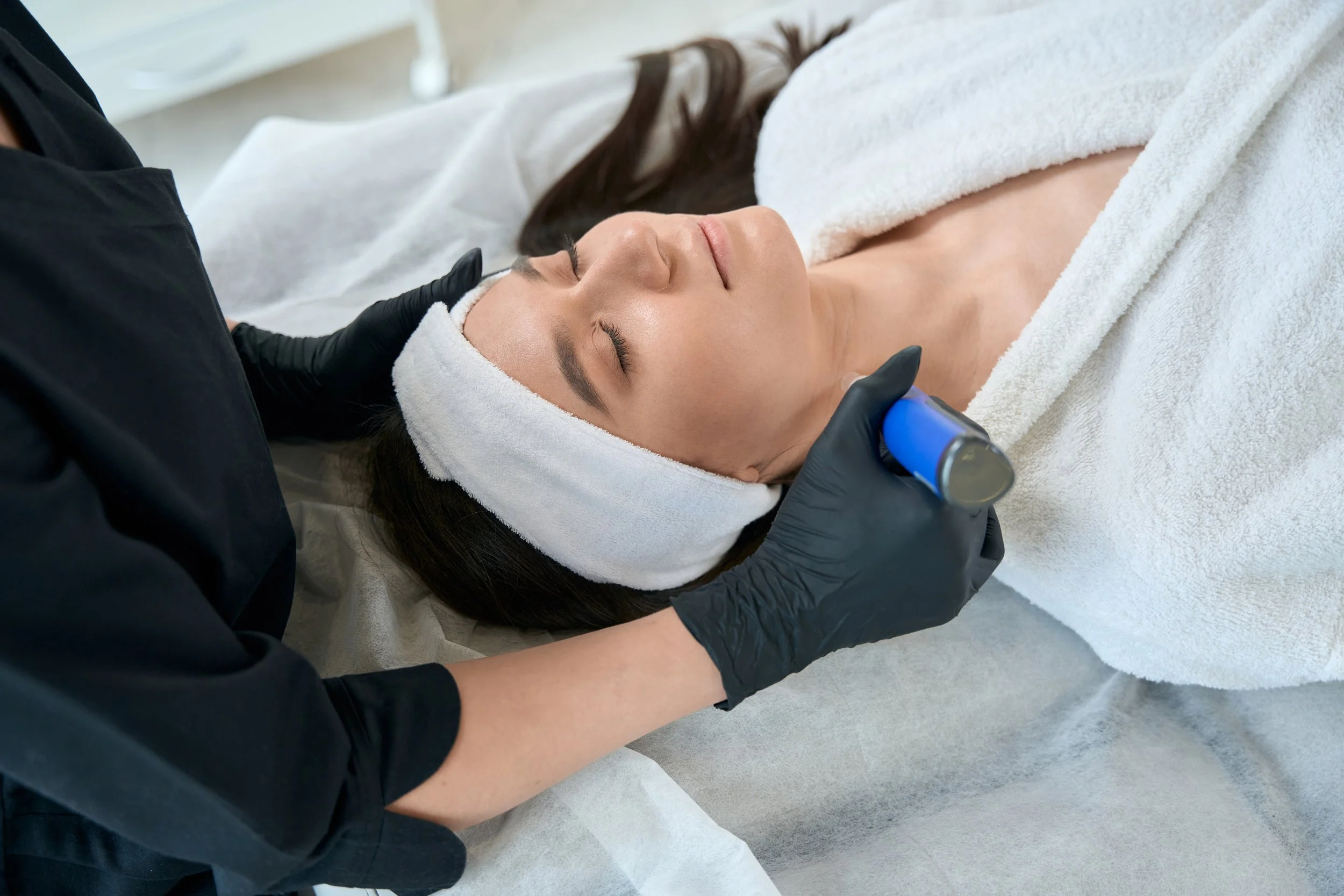 Woman receiving a facial treatment with a device, wearing a headband, therapist with gloves.
