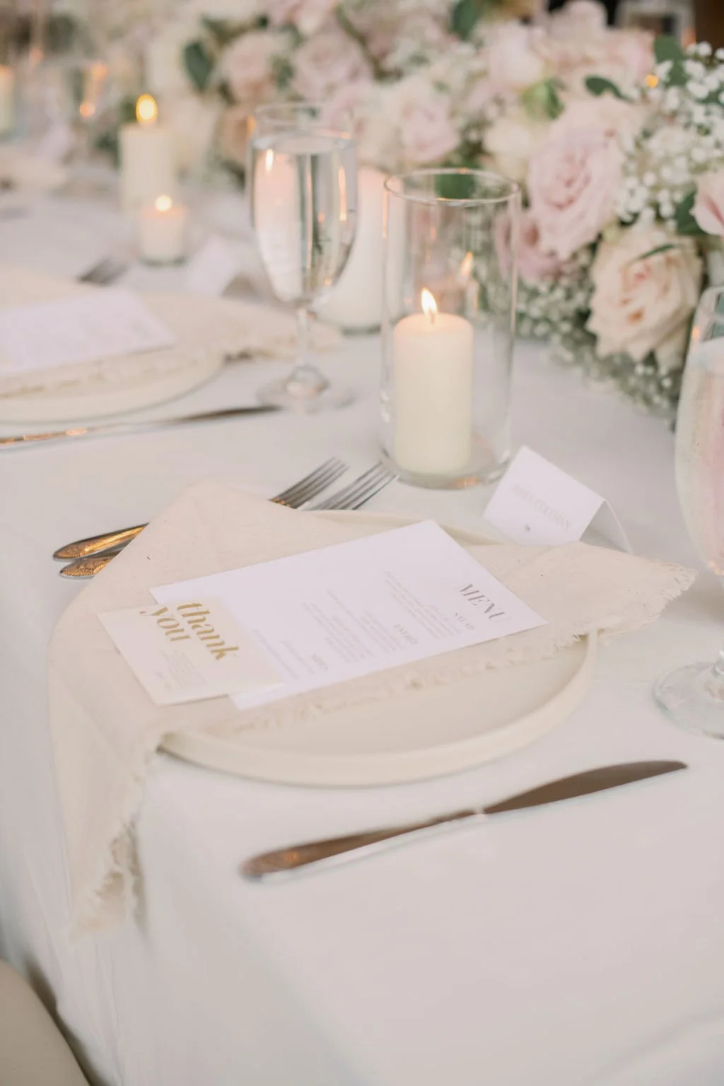 Elegant wedding table setting with white plates, menus, thank you cards, a lit candle, wine glasses, and pink floral arrangements.
