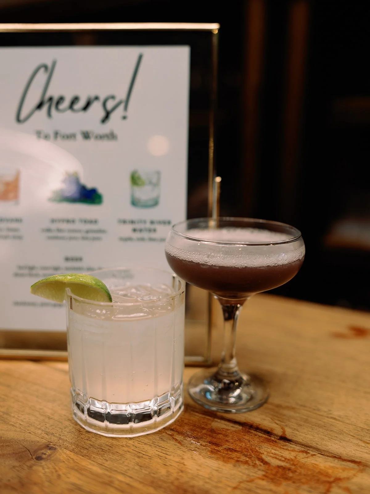 Two cocktails on a wooden table with a framed menu in the background. One is a clear drink with a lime wedge, served in a rocks glass. The other is a darker drink in a coupe glass.