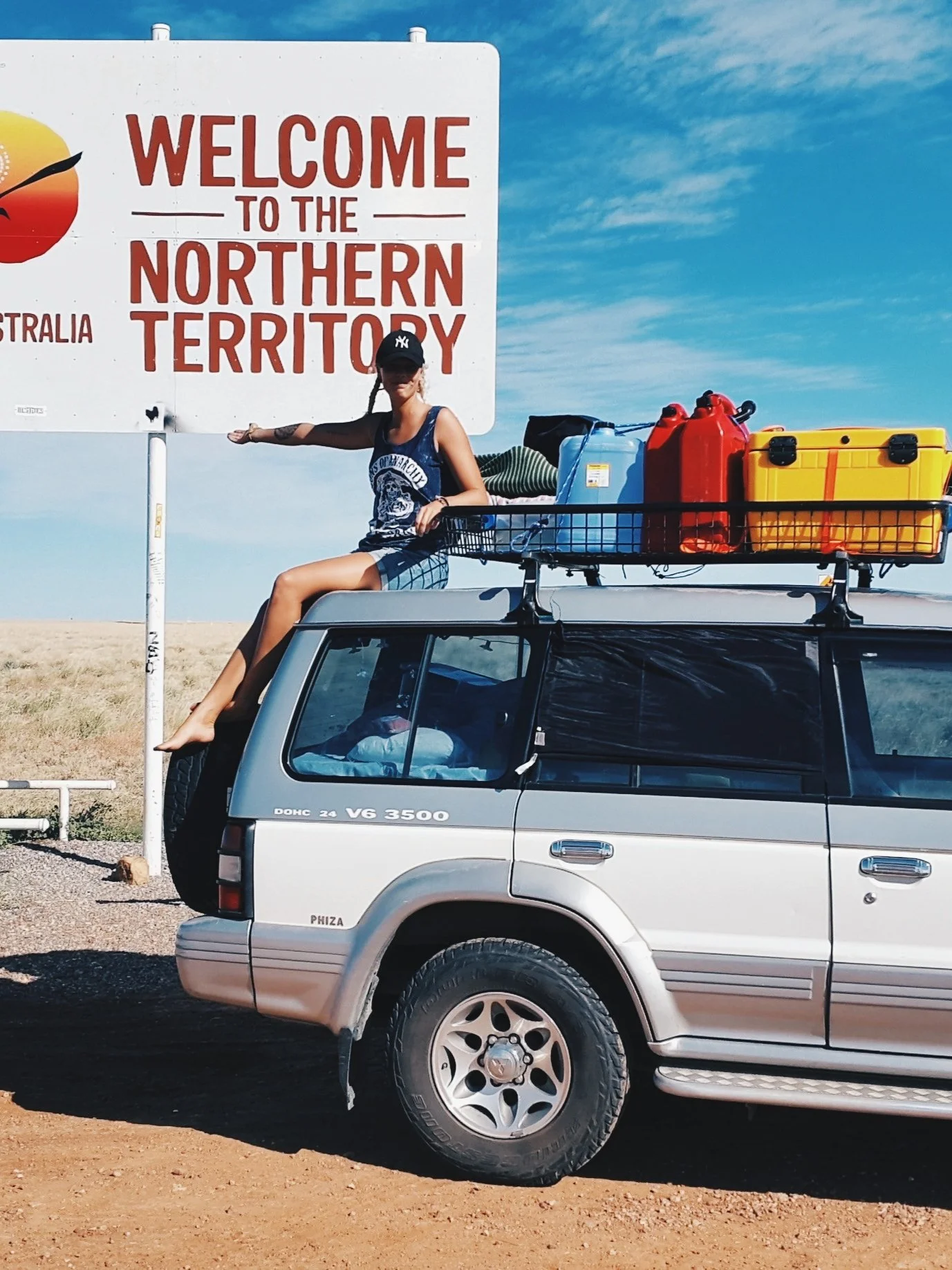 Johanna sitter på taket på sin van i Australien och pekar på en skylt som det står "Welcome to thhe Northern Territory" på.