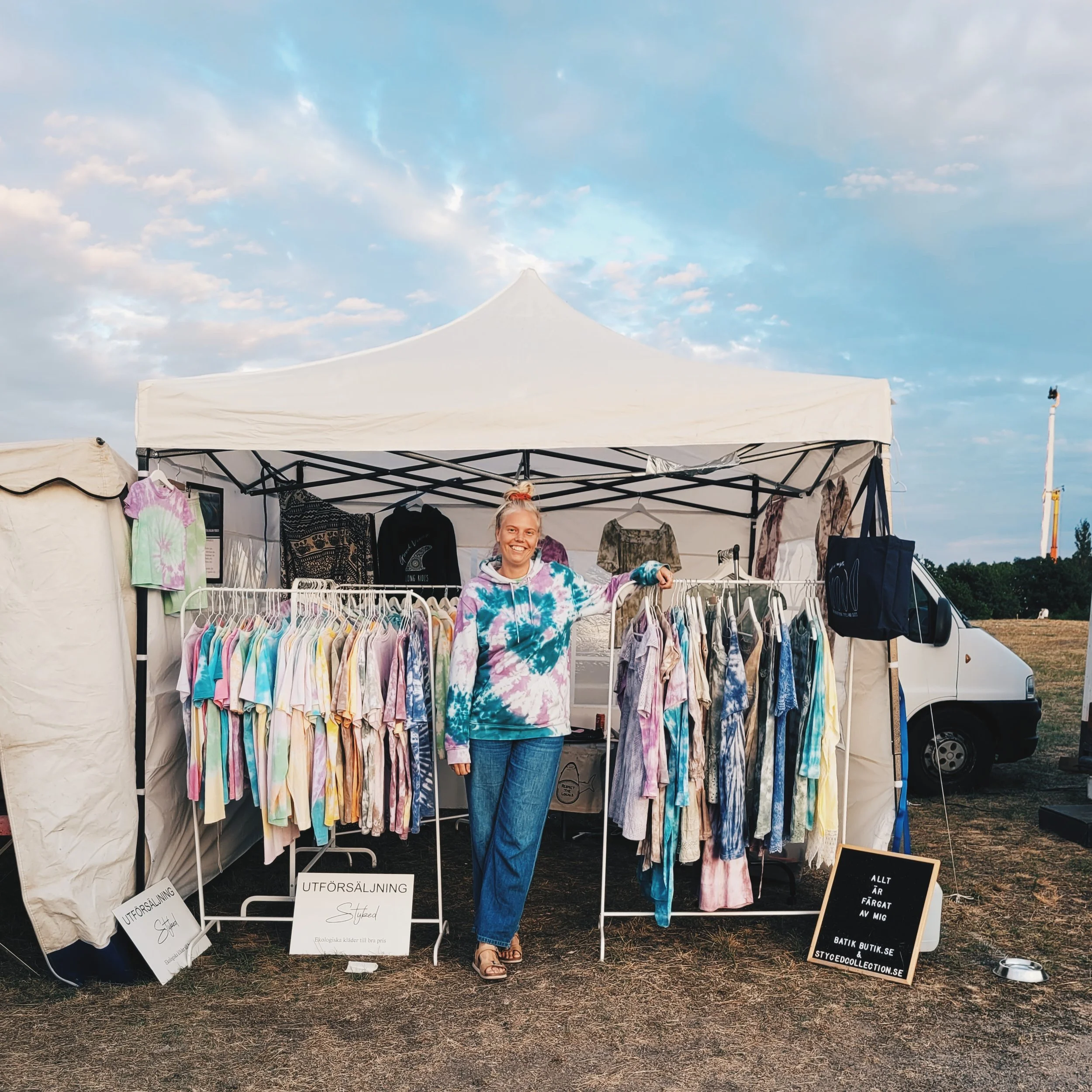 Johanna står och poserar vid sitt tält på en av de marknader hon säljer på. Vid sin sida har hon massor med färgglada batikkläder och i bakgrunden syns hennes van..