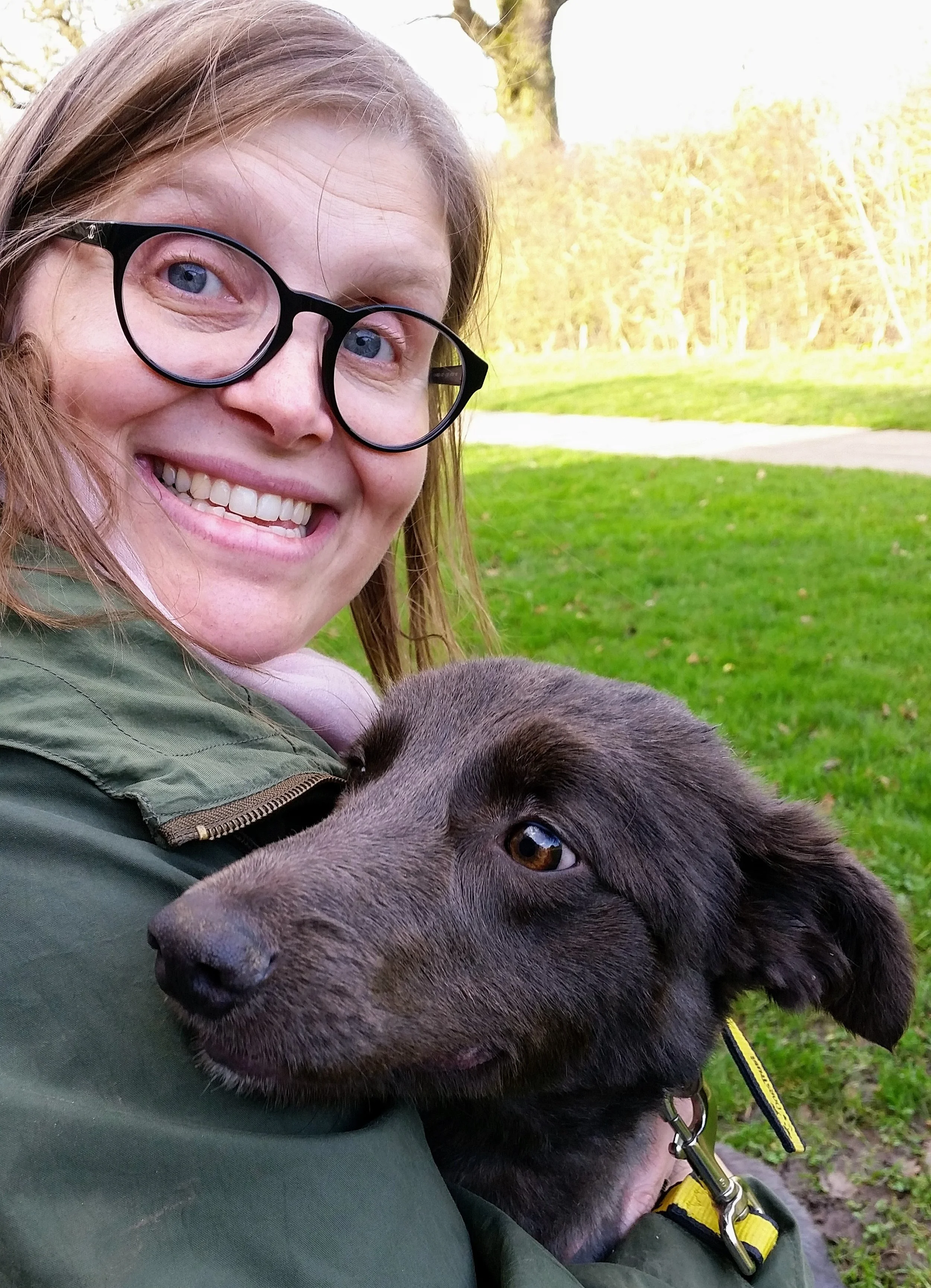 A woman with glasses smiling and holding a gray puppy outdoors on a grassy area with trees in the background.