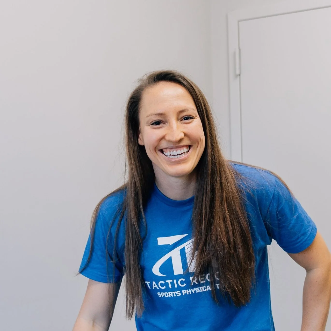 Woman with long brown hair smiling, wearing a blue T-shirt with white text about tactics, sports, and physical therapy, standing against a plain wall.