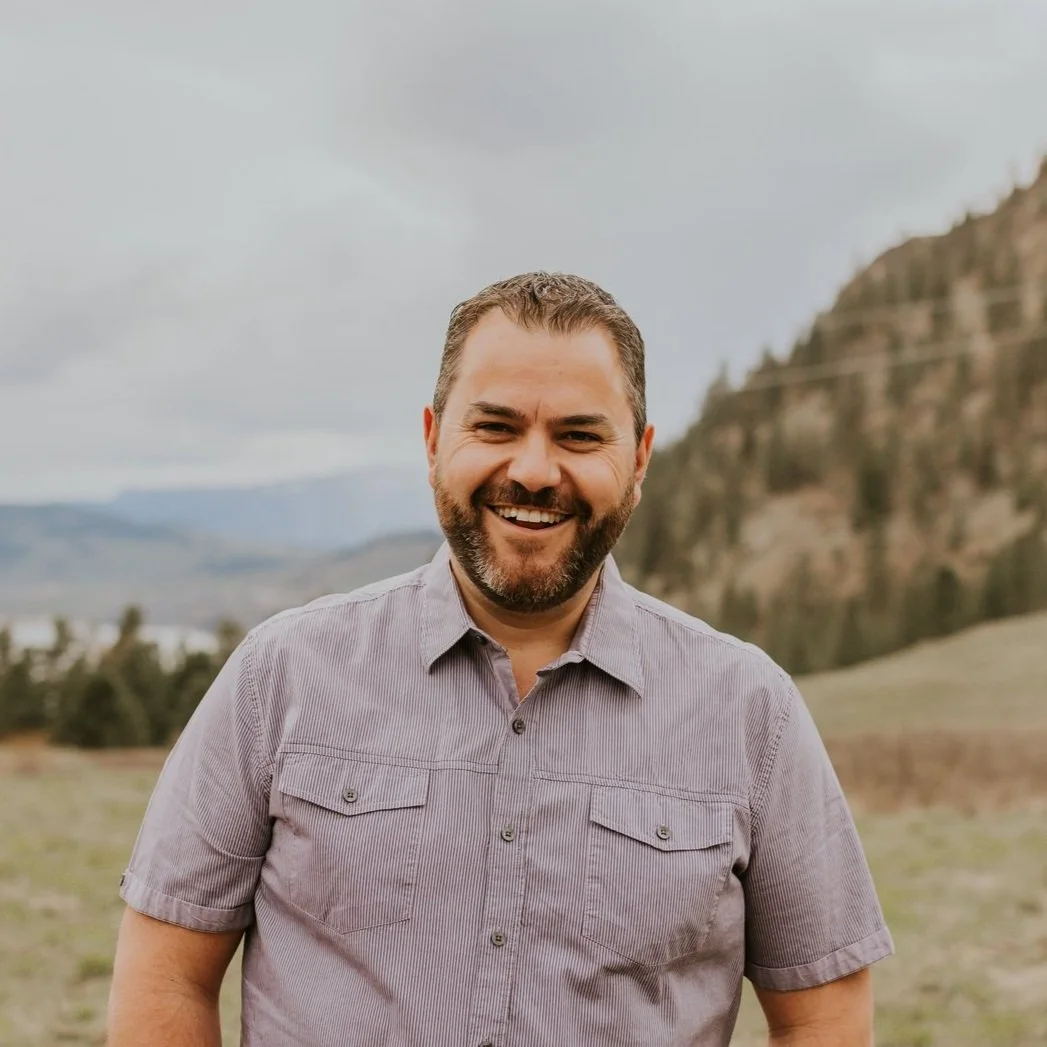 A man with a beard and short hair smiling outdoors with mountains and cloudy sky in the background.