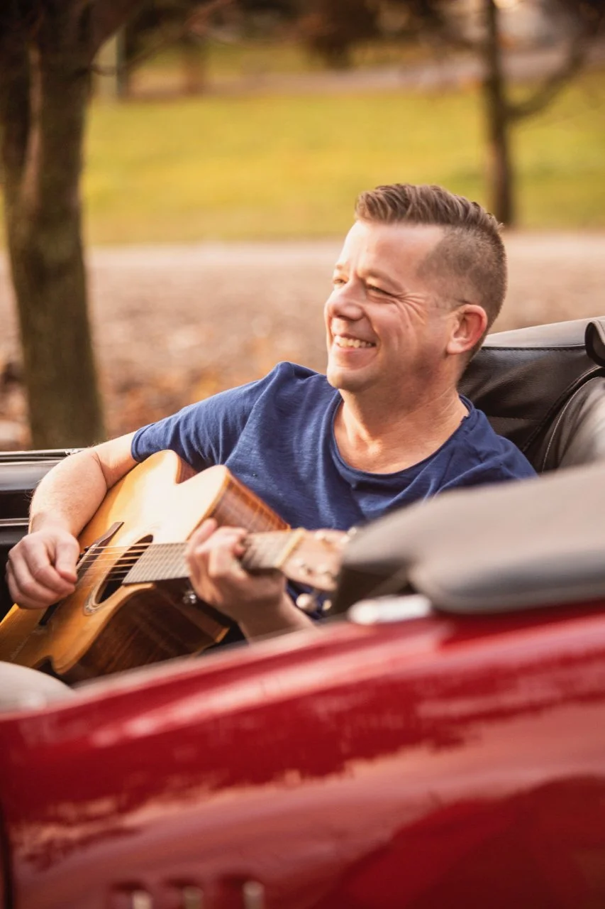 A man with short hair and a blue shirt playing an acoustic guitar and smiling while sitting in the driver’s seat of an open-top car outdoors.