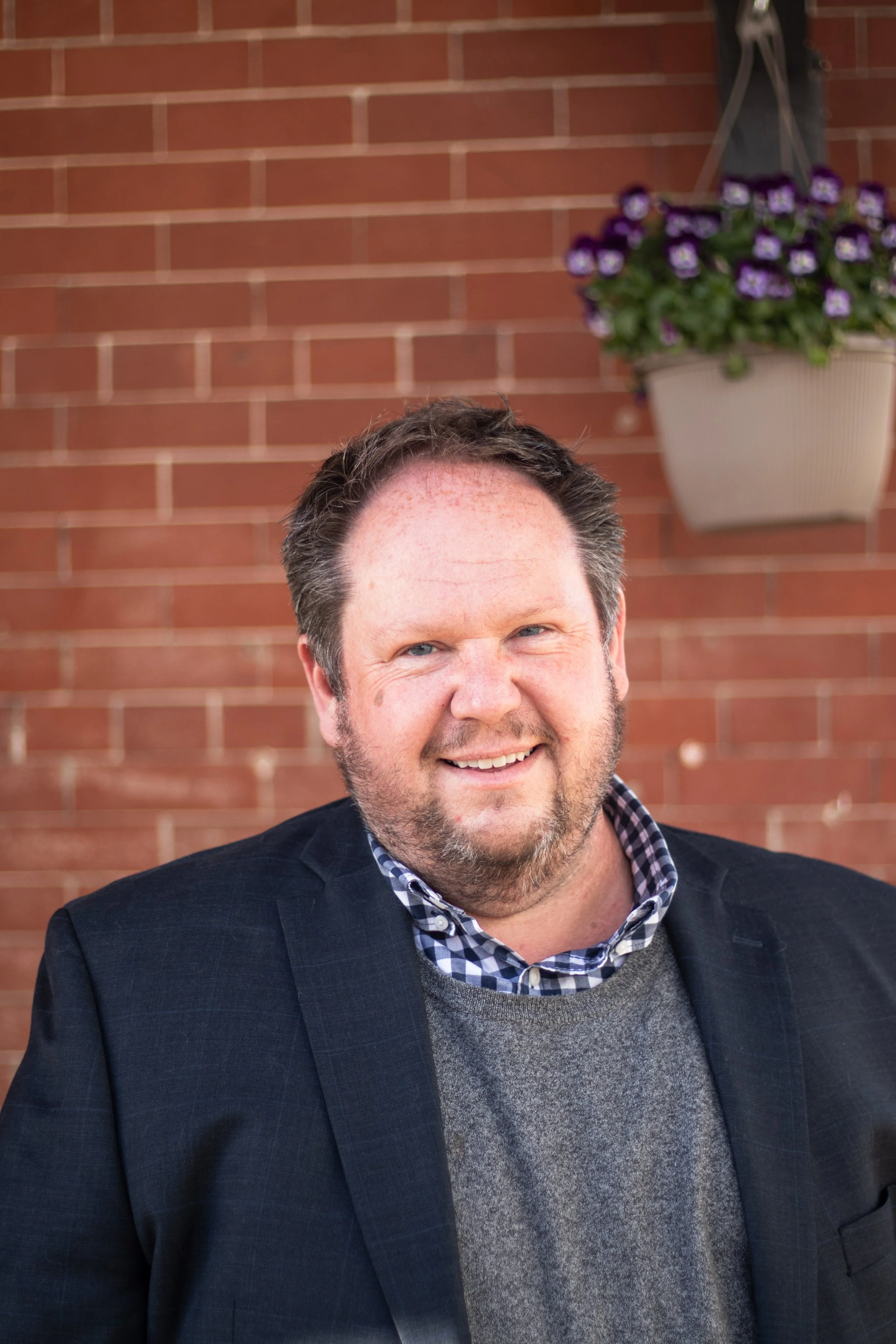 A smiling man with short brown hair and a beard, wearing a dark blazer, plaid shirt, and sweater, standing outdoors in front of a brick wall and a hanging flower pot with purple flowers.