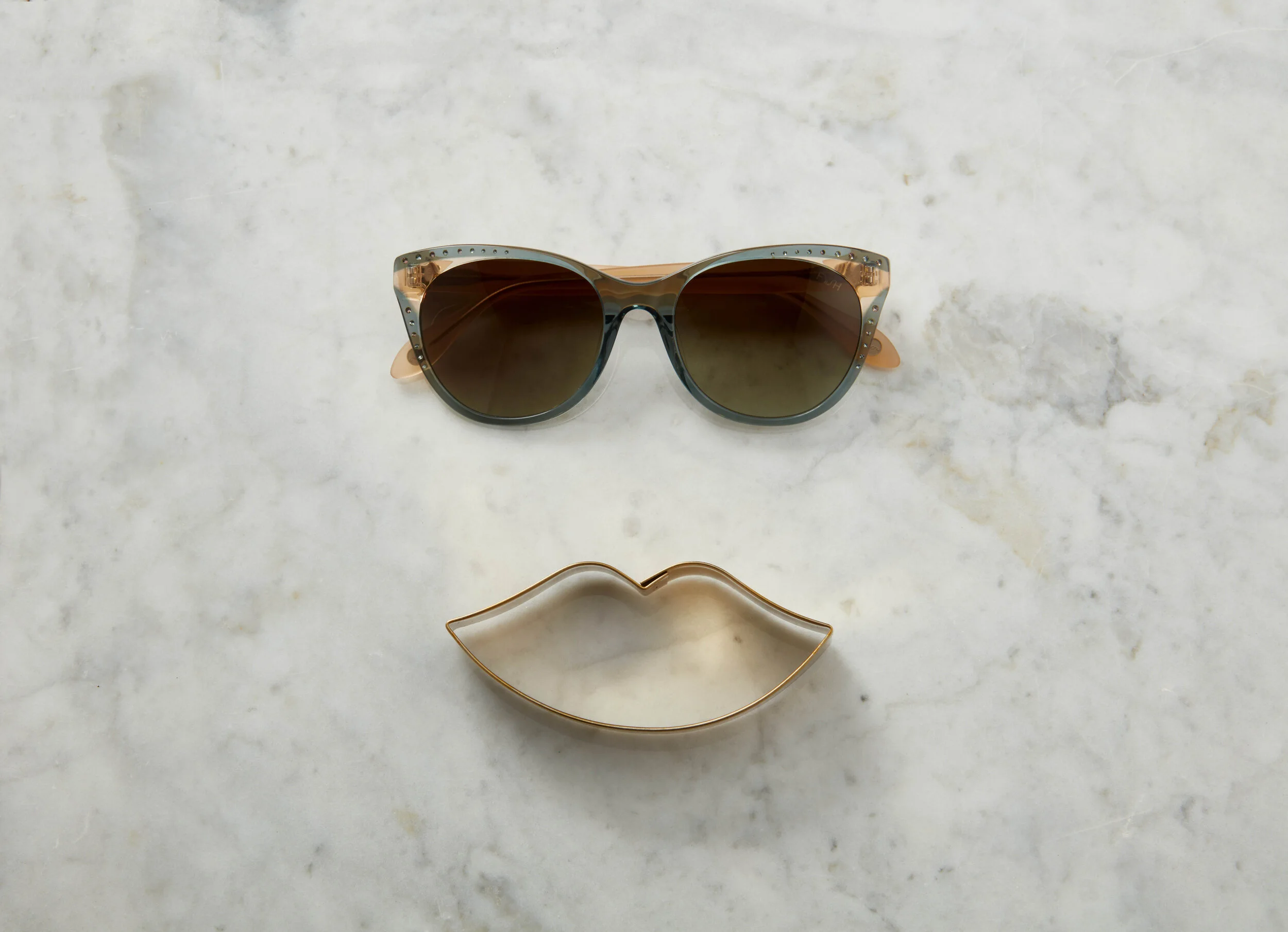 A pair of sunglasses with dark lenses positioned above a metallic cookie cutter in the shape of lips on a white marble surface.