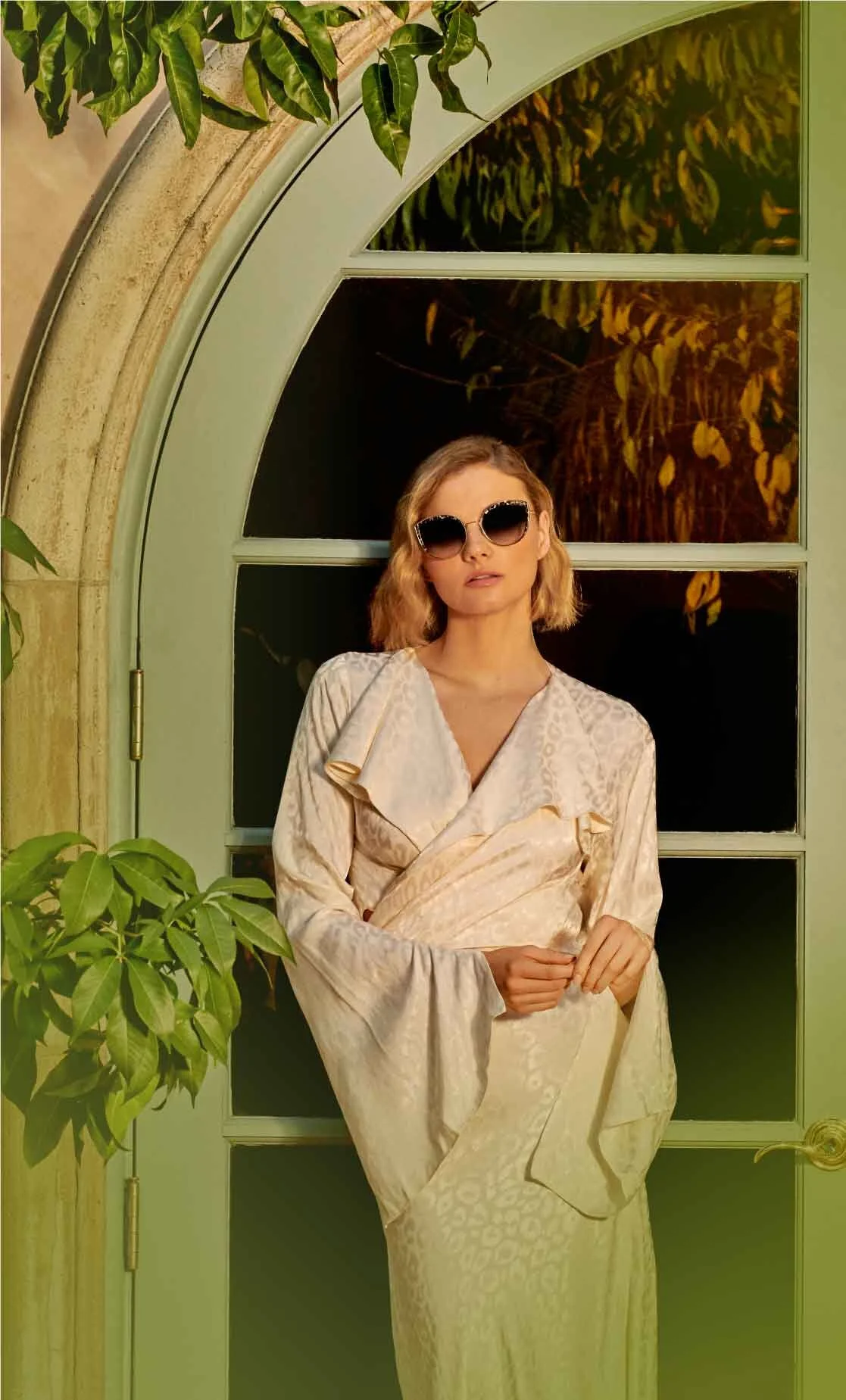 A woman wearing large black sunglasses and a beige satin dress standing in front of a green arched door with glass panes. There are green plants on the side.