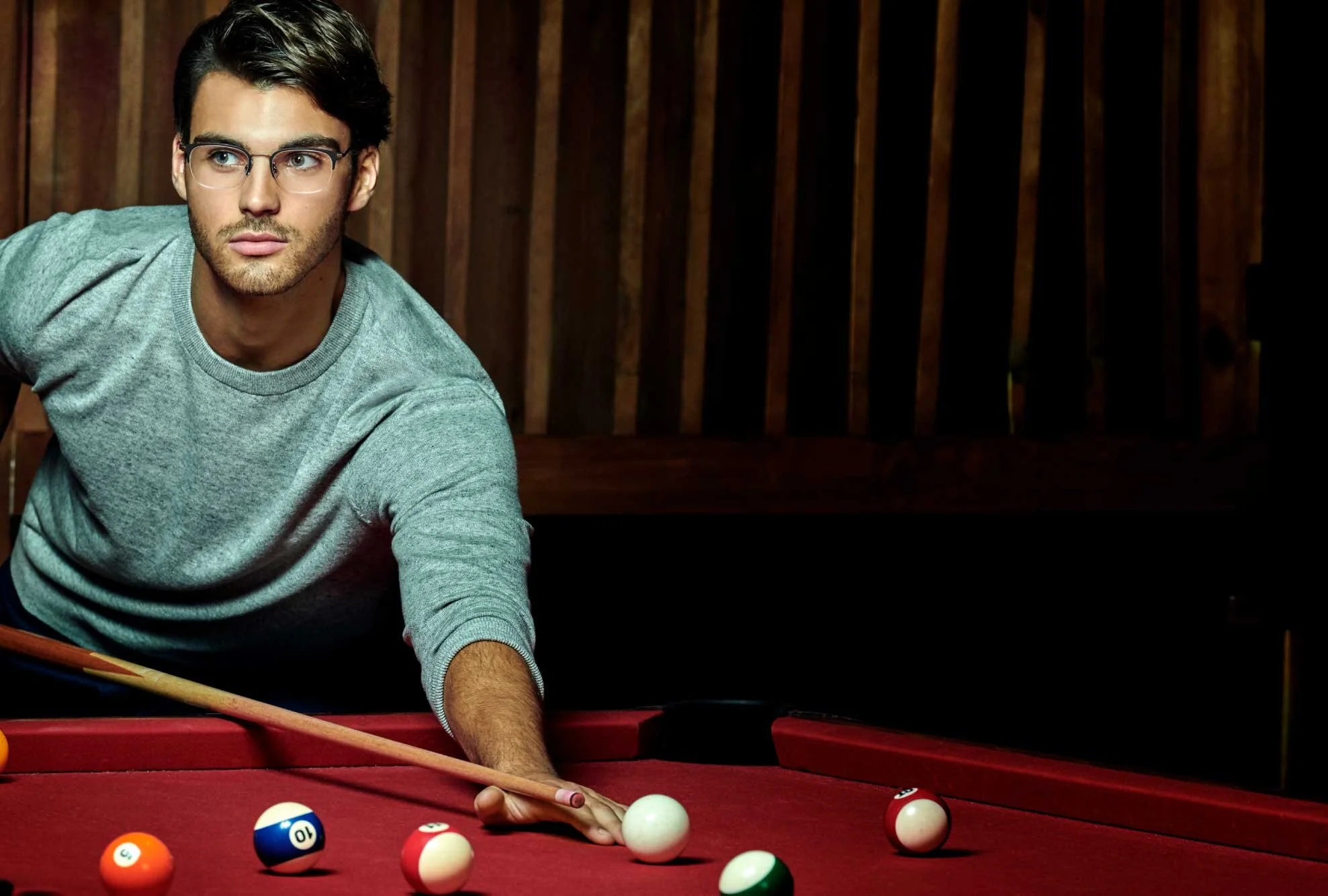 A young man with glasses, wearing a gray sweatshirt, leaning over a red pool table, preparing to take a shot with a cue stick. Colored billiard balls are scattered on the table, and a wooden wall is in the background.