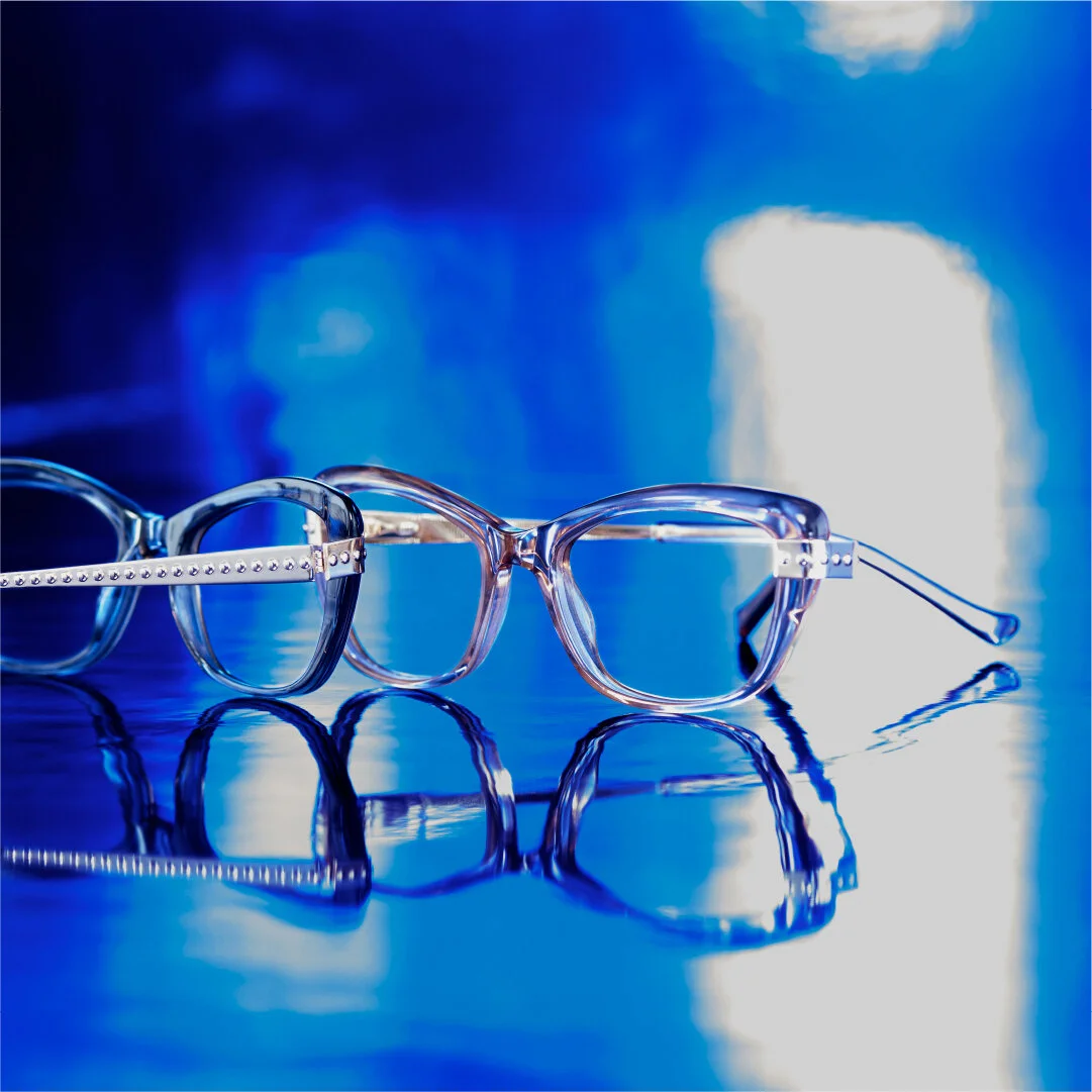 Two pairs of clear eyeglasses with reflections on a shiny, reflective surface, illuminated by blue and white lighting.