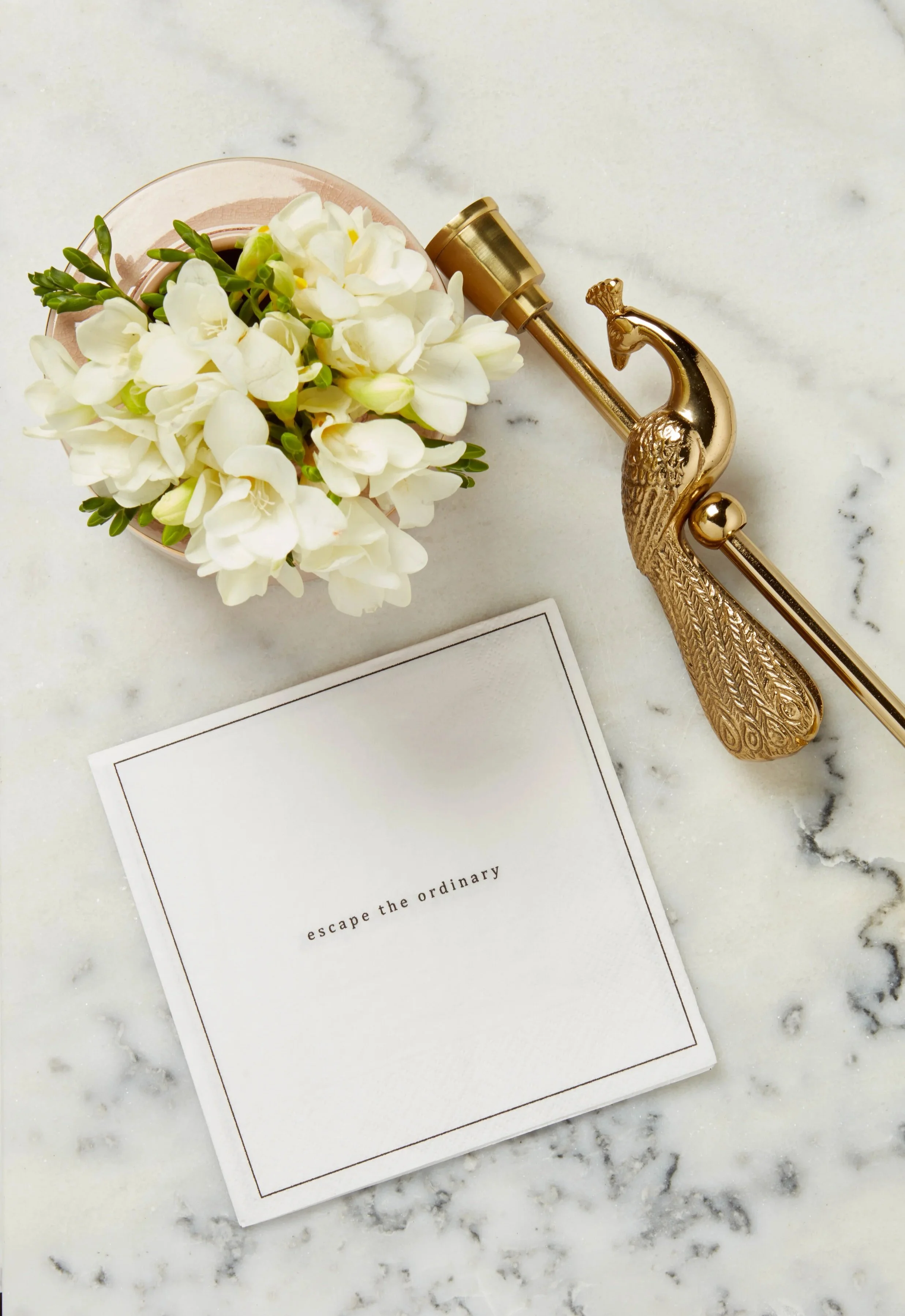 A flat lay of a gold peacock-shaped eyelash curler, a small pink pot with white flowers, and a white card with the text "escape the ordinary" on a marble surface.