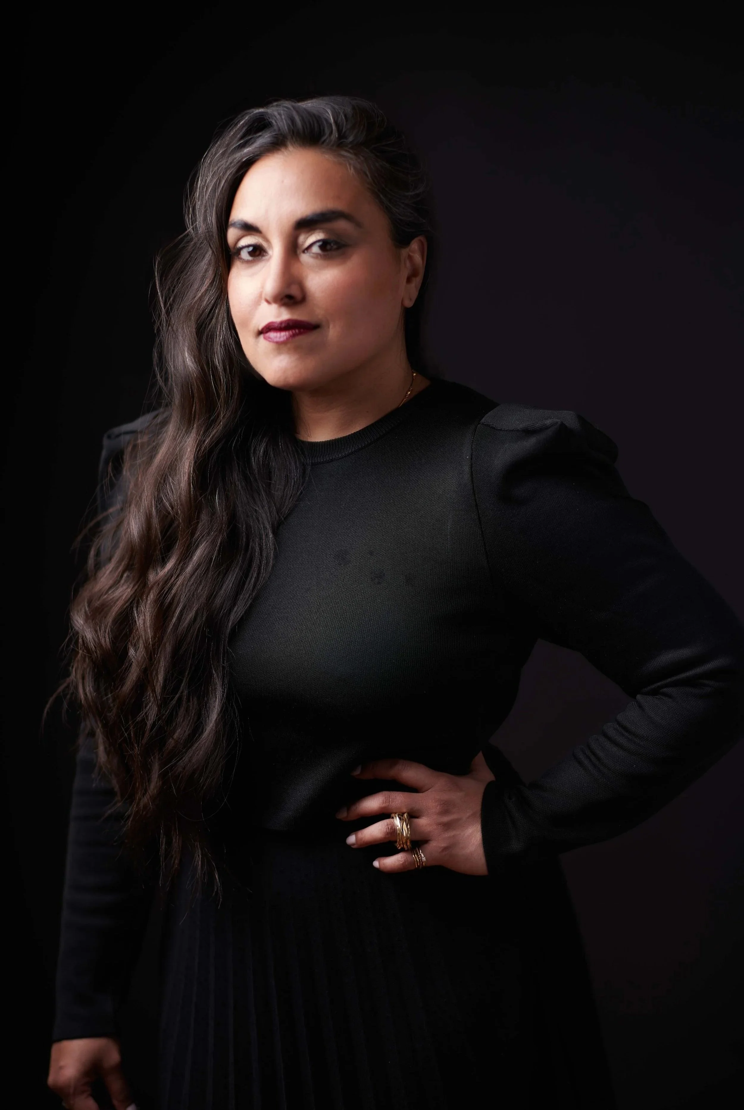A woman with long, wavy dark hair wearing a black long-sleeve top with puffed shoulders, standing against a dark background.