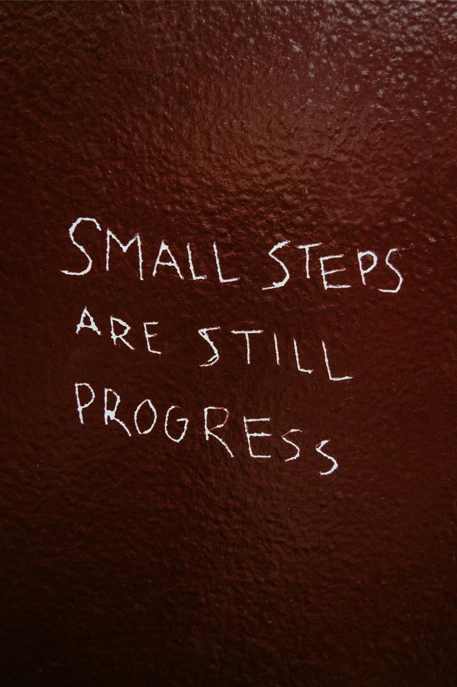 White handwritten text on a brown textured wall reads, 'Small steps are still progress.'