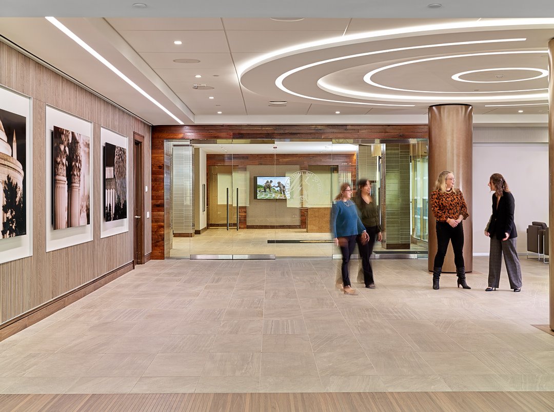 A modern, well-lit lobby with three women conversing near a large column and a glass wall. The background features a hallway with art on the walls and a television screen.