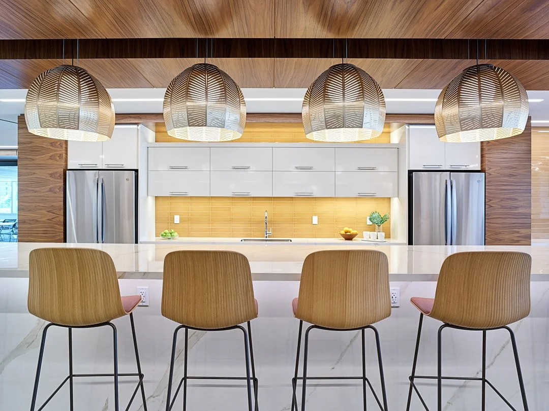 Modern kitchen with four wooden bar stools at a white marble counter, stainless steel refrigerators, white cabinetry, yellow backsplash, pendant lights, and minimal decor.