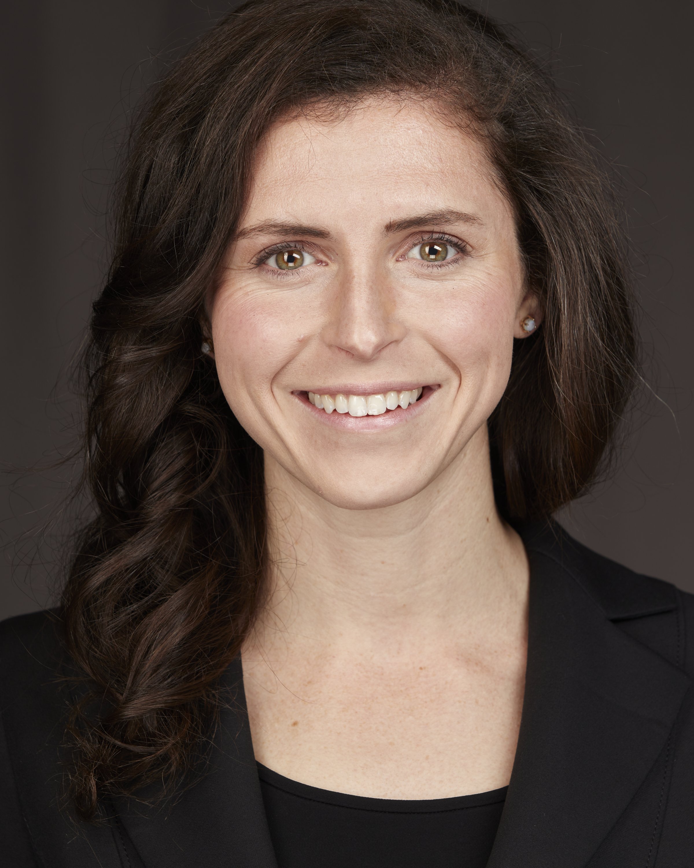 Close-up portrait of a smiling woman with brown hair, hazel eyes, and pearl earrings, wearing a black top and blazer against a dark background.
