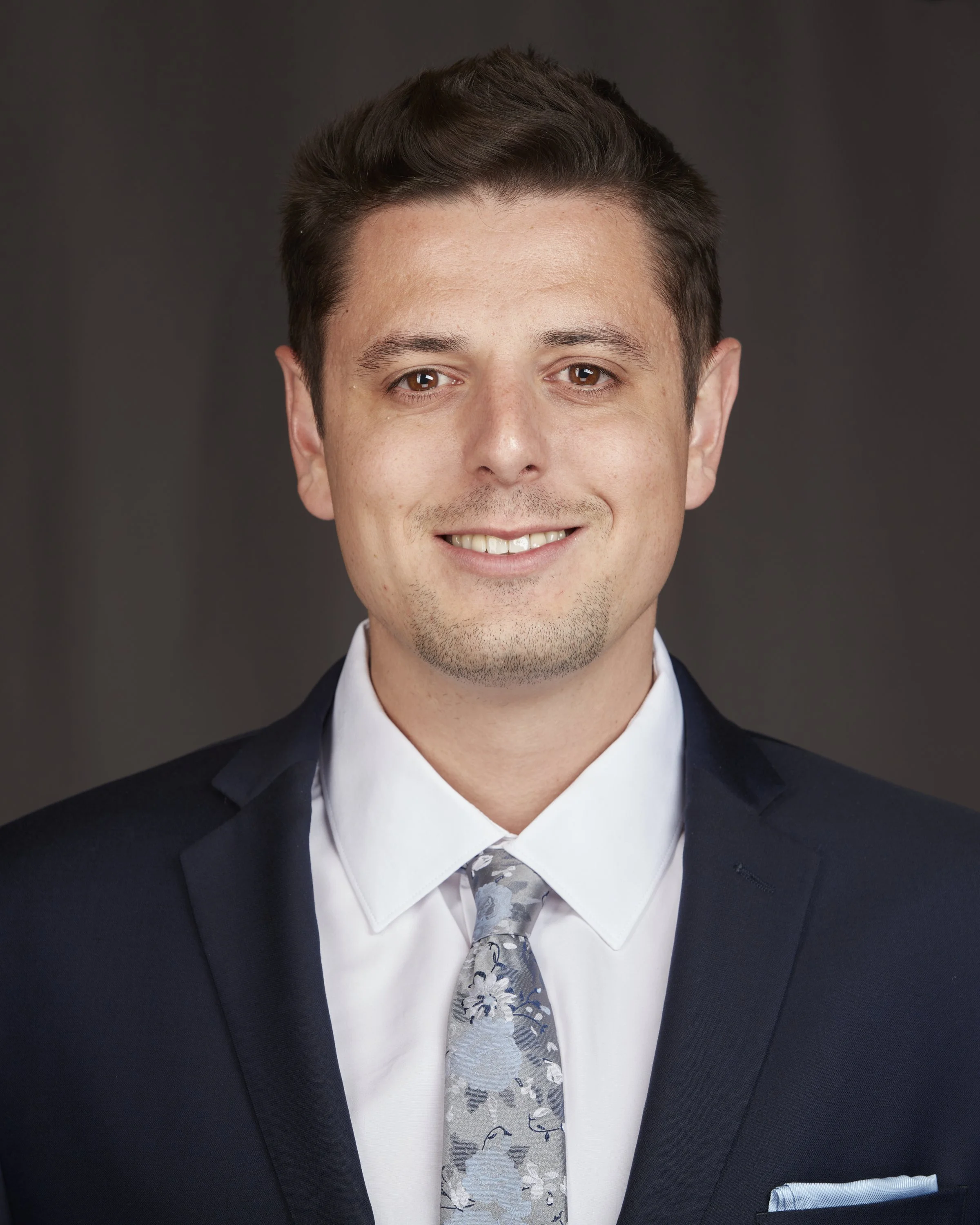 Close-up of a young man smiling, dressed in a navy suit, white shirt, and a floral patterned tie, against a dark gray background.