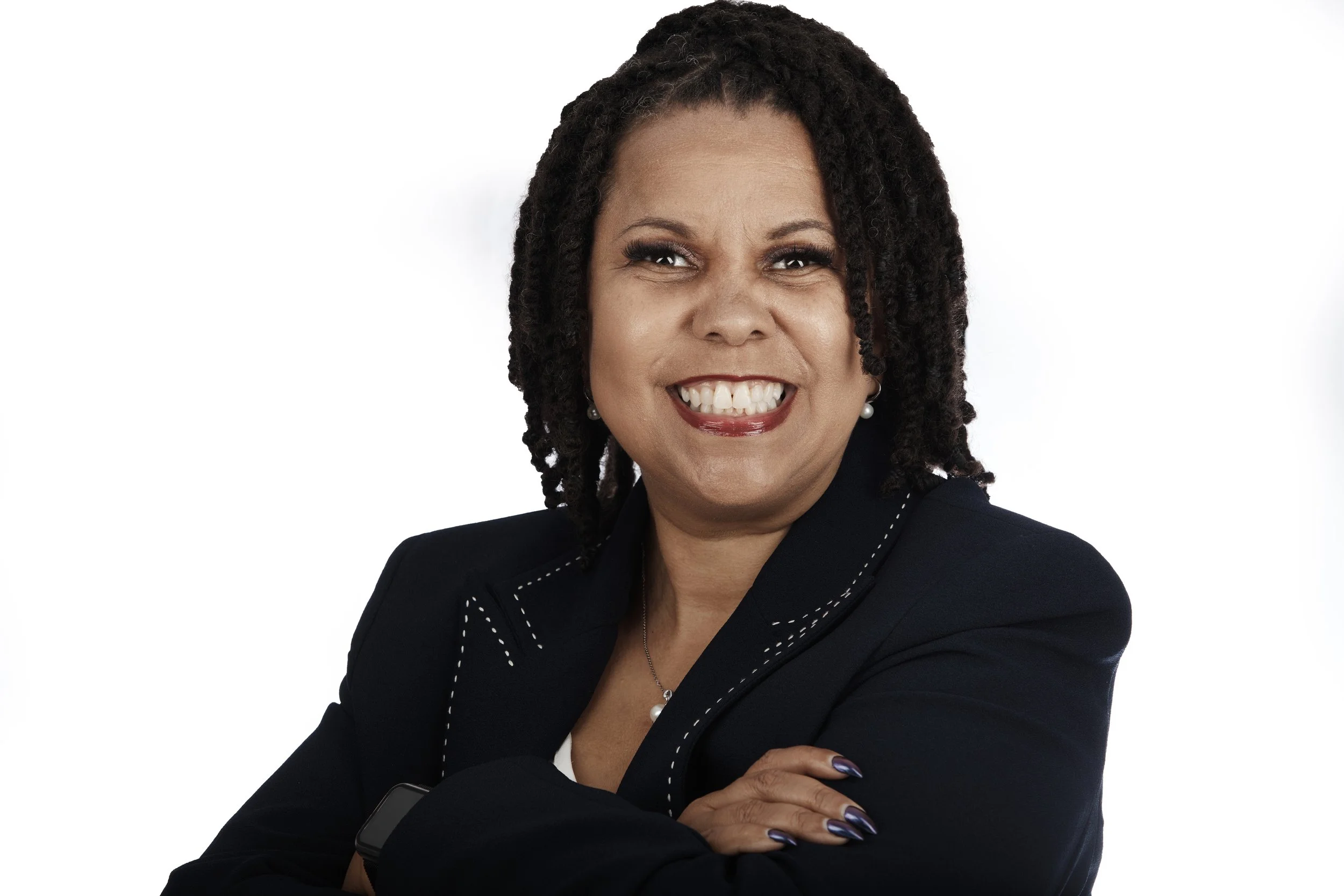 A smiling woman with dark, textured hair styled in dreadlocks, wearing a dark blazer with white stitching, earrings, and makeup with purple lipstick, posing against a white background.