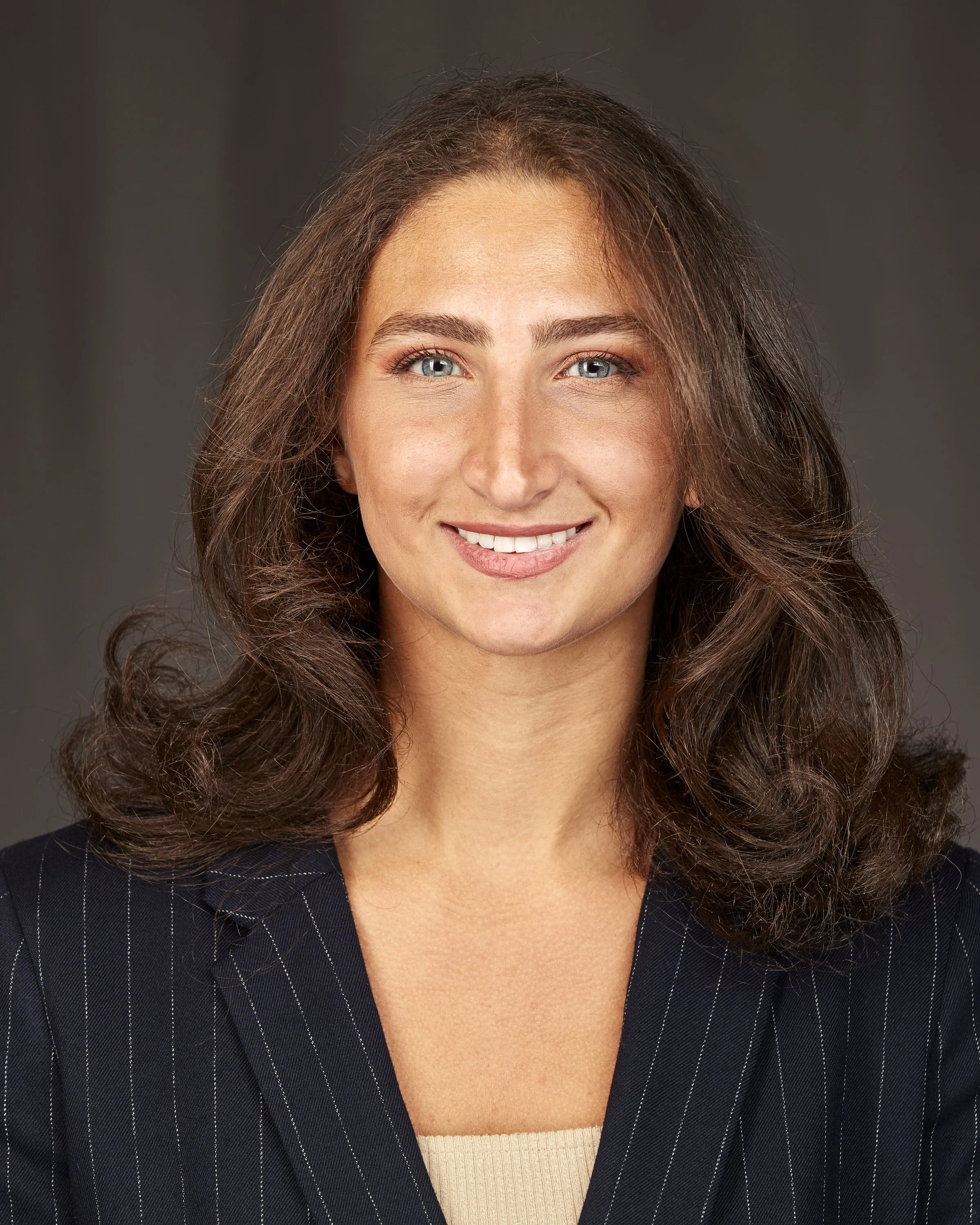 Close-up of a smiling woman with wavy brown hair, wearing a black pinstripe suit jacket and a beige top, against a dark background.