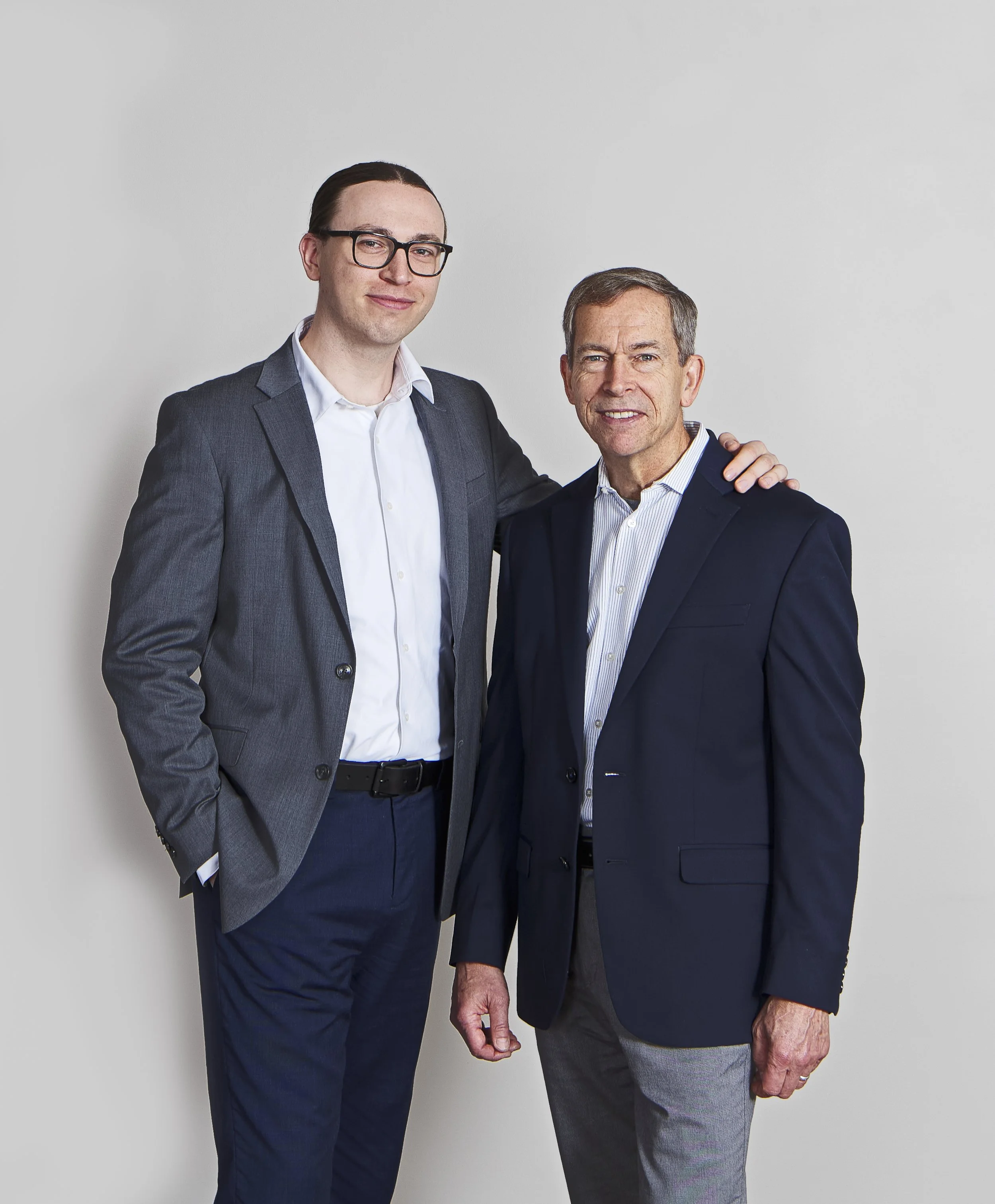 Two men standing together, one younger with glasses and longer hair, in a suit, the other older with shorter hair, in a blazer, with the younger man's arm around the older man's shoulder, against a plain white background.