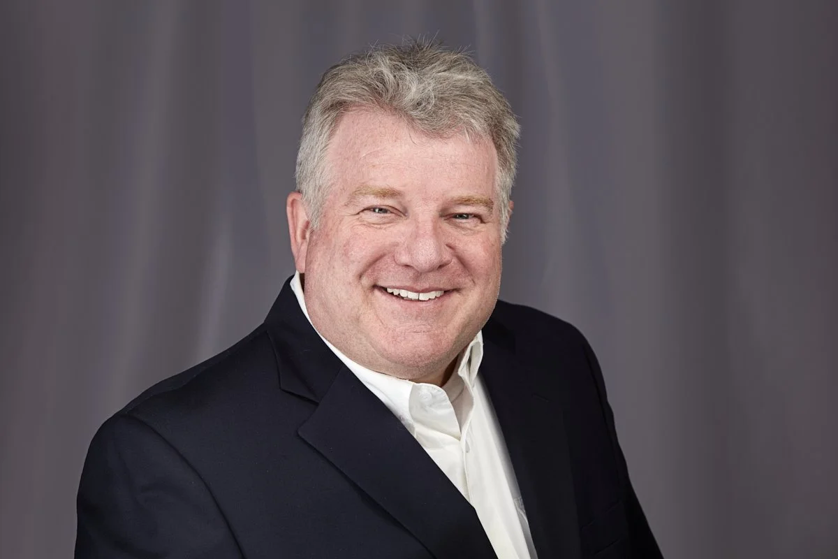 Smiling man wearing a black suit jacket and white shirt in front of a plain gray background.