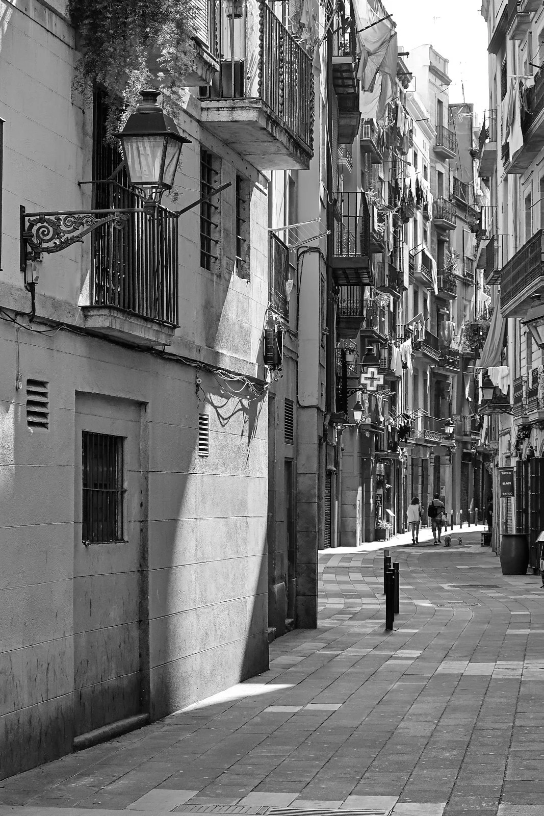 A narrow urban street scene in black and white with multi-story buildings, balconies, laundry hanging outside, street lamps, and two people walking with a dog.