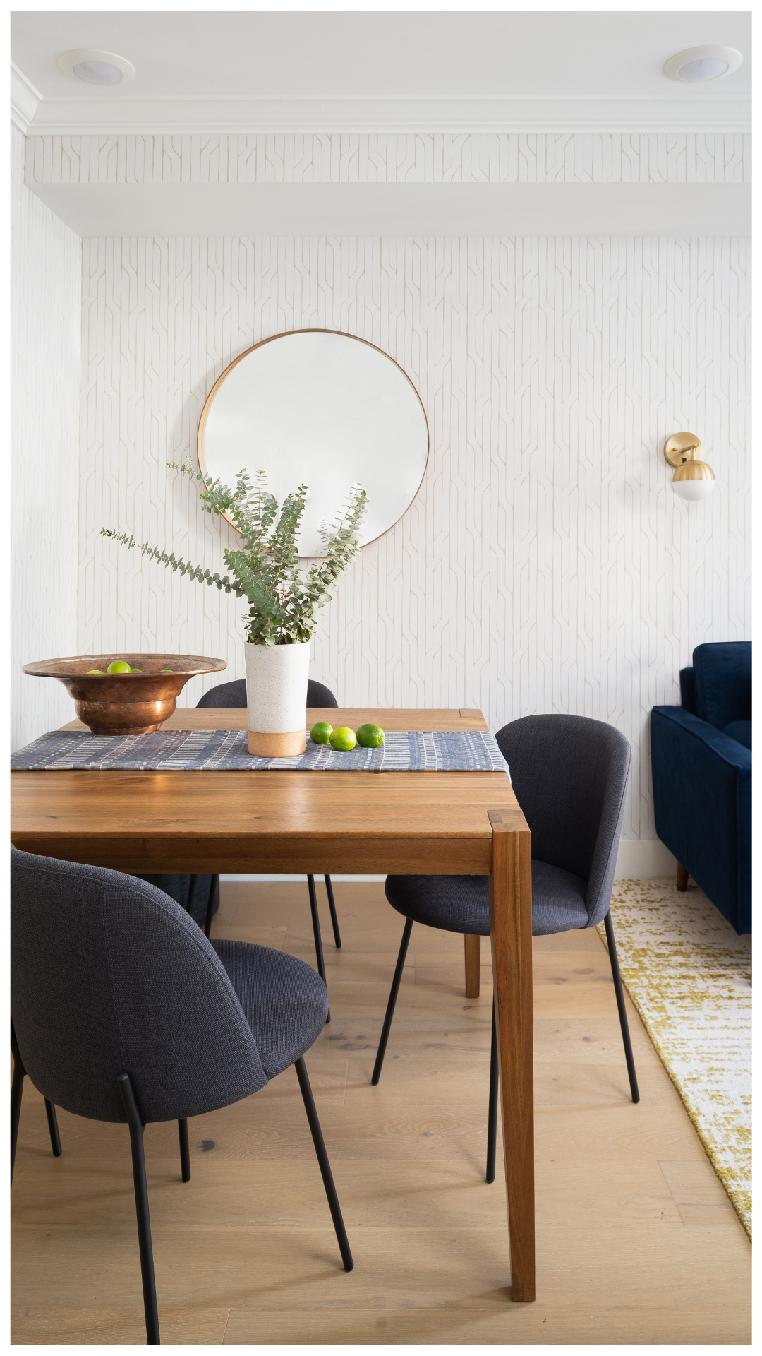 A cohesive open-concept living and dining orchestration featuring a blue sectional, mid-century modern wood dining table, and geometric wallpaper, designed for clear spatial definition and functional flow.