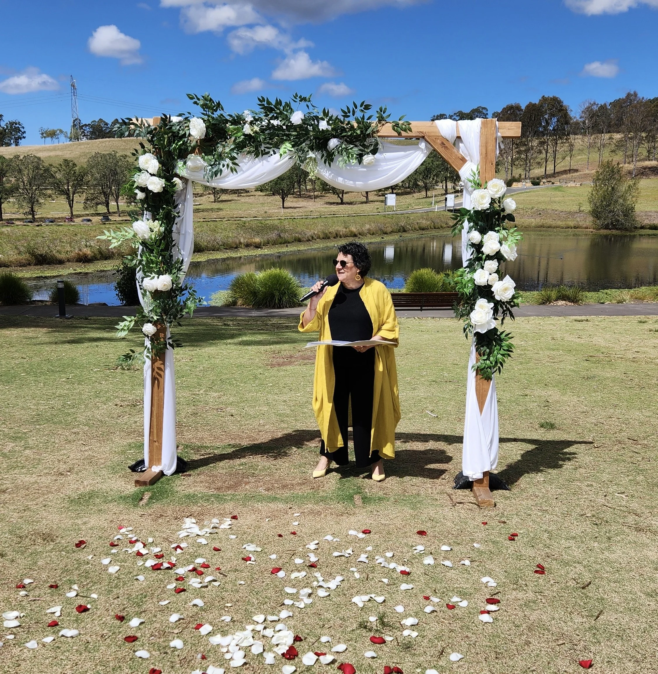 Person in yellow coat speaking under floral wedding arch outdoors on a sunny day