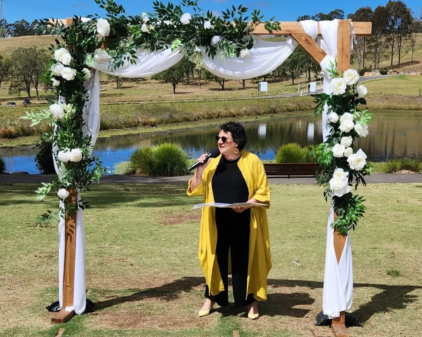 Person in yellow coat speaking under floral wedding arch outdoors on a sunny day
