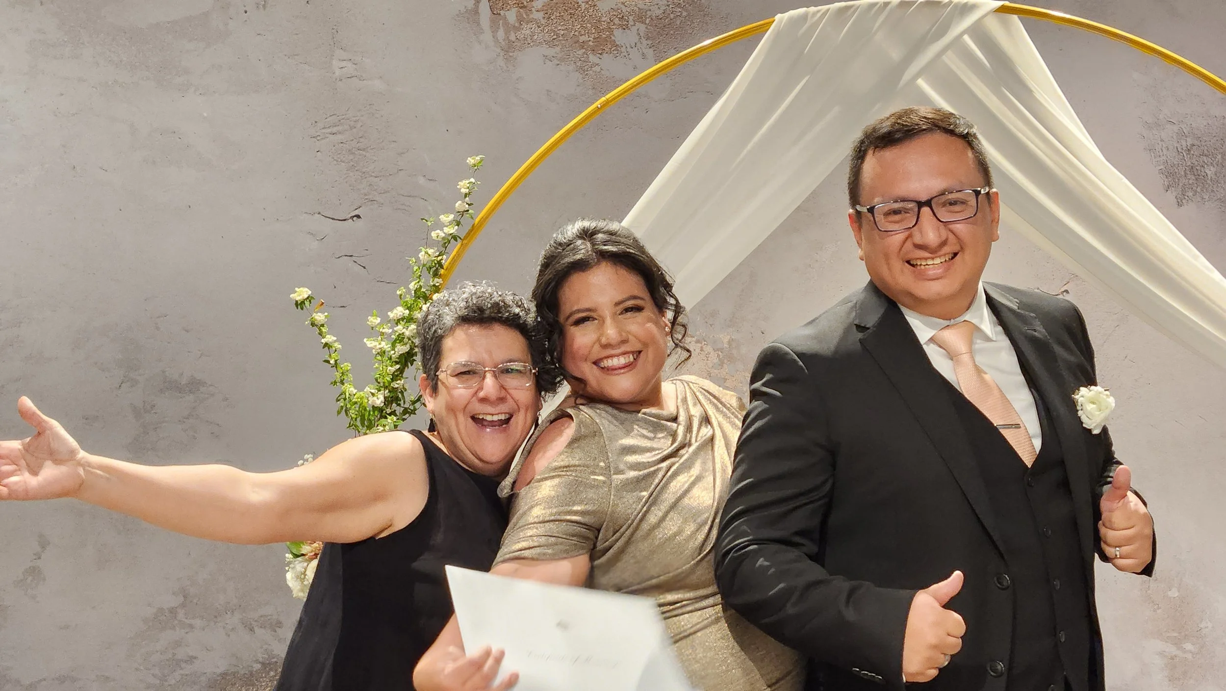 Three people posing and smiling at an event with floral and fabric backdrop.