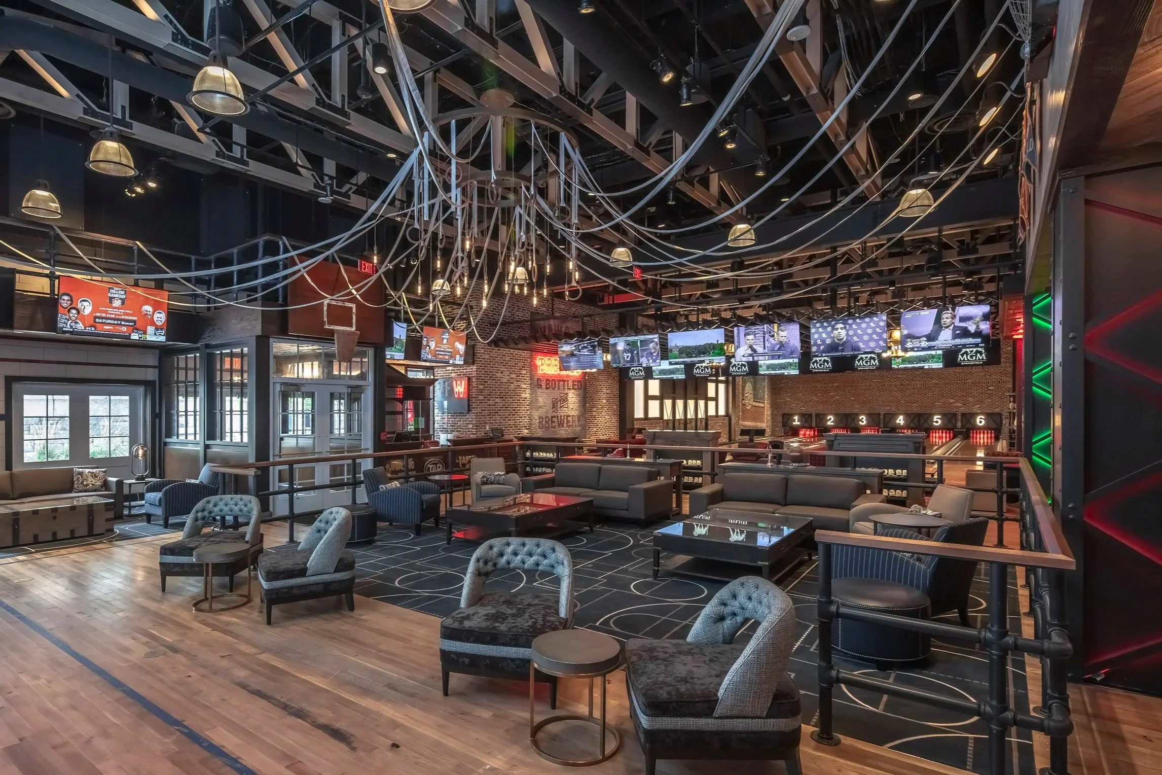 A spacious sports bar with multiple wall-mounted televisions displaying various sports events, comfortable seating areas with armchairs and couches, and decorative ceiling ropes and lighting fixtures.