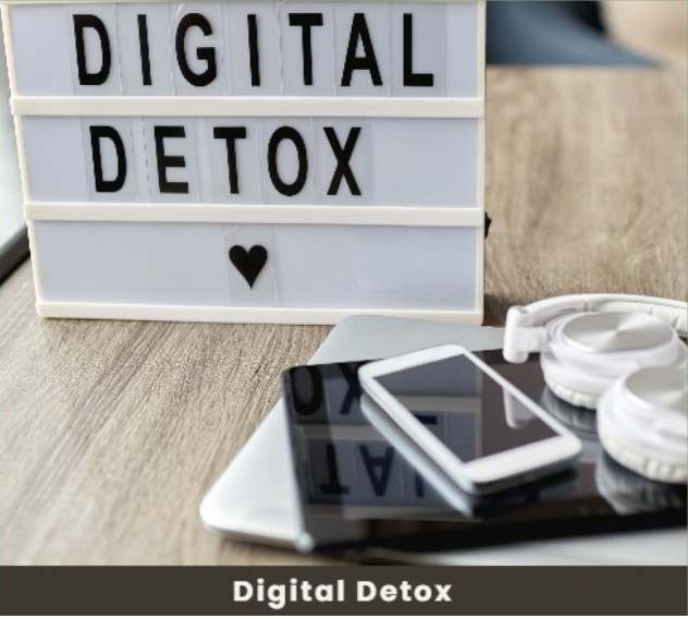 A white letter board with the words 'Digital Detox' and a black heart, placed on a wooden surface alongside a smartphone, a tablet, and a pair of white headphones.