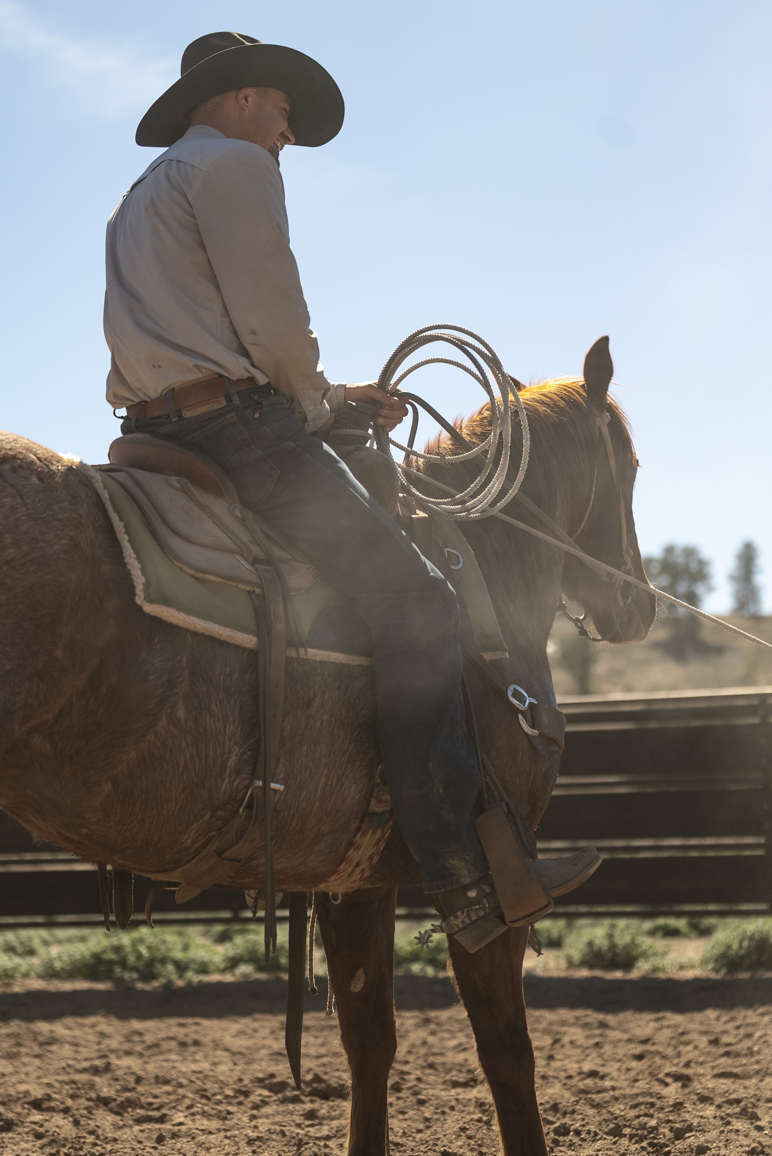 A man wearing a cowboy hat and sunglasses riding a brown horse through a grassy field with trees in the background during sunset.