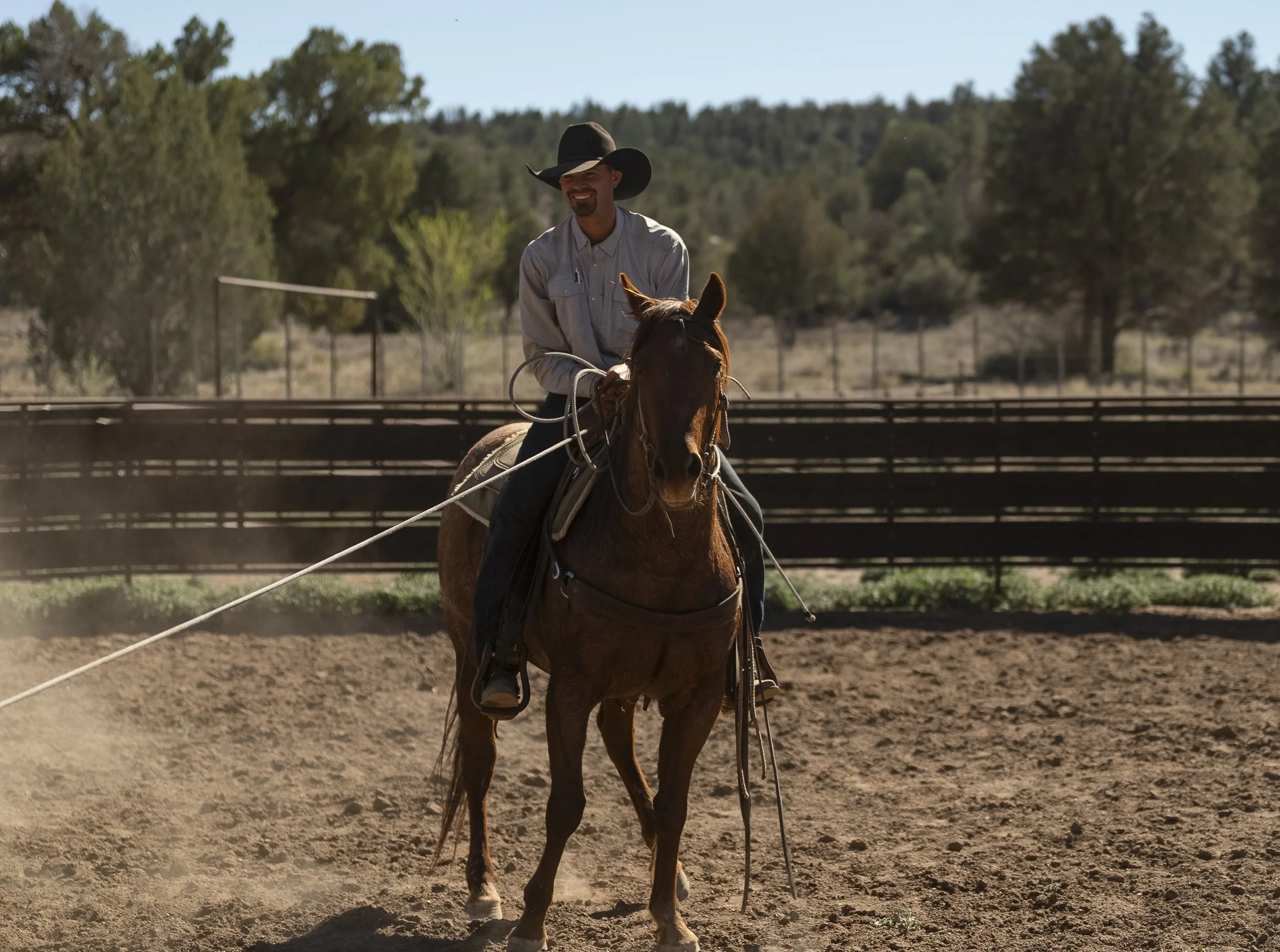 A man wearing a cowboy hat and sunglasses riding a brown horse through a grassy field with trees in the background during sunset.