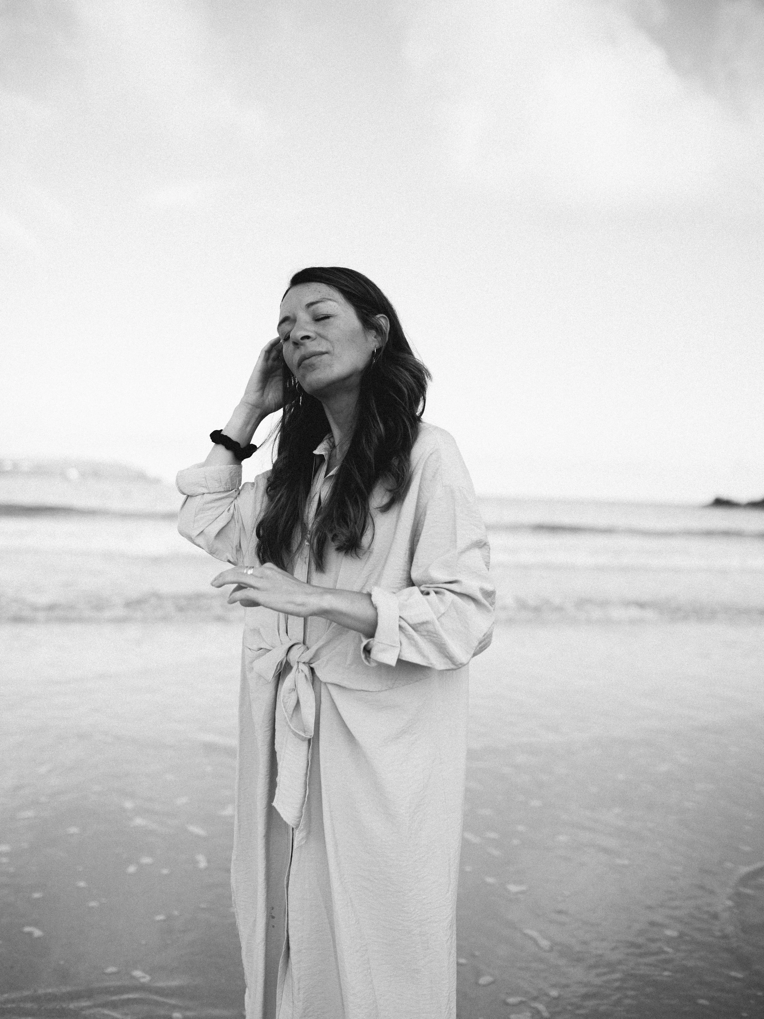 A woman with long dark hair standing on a beach, dressed in a light-colored, long-sleeve dress with a tied waist, with her eyes closed and hand touching her face, in black and white.