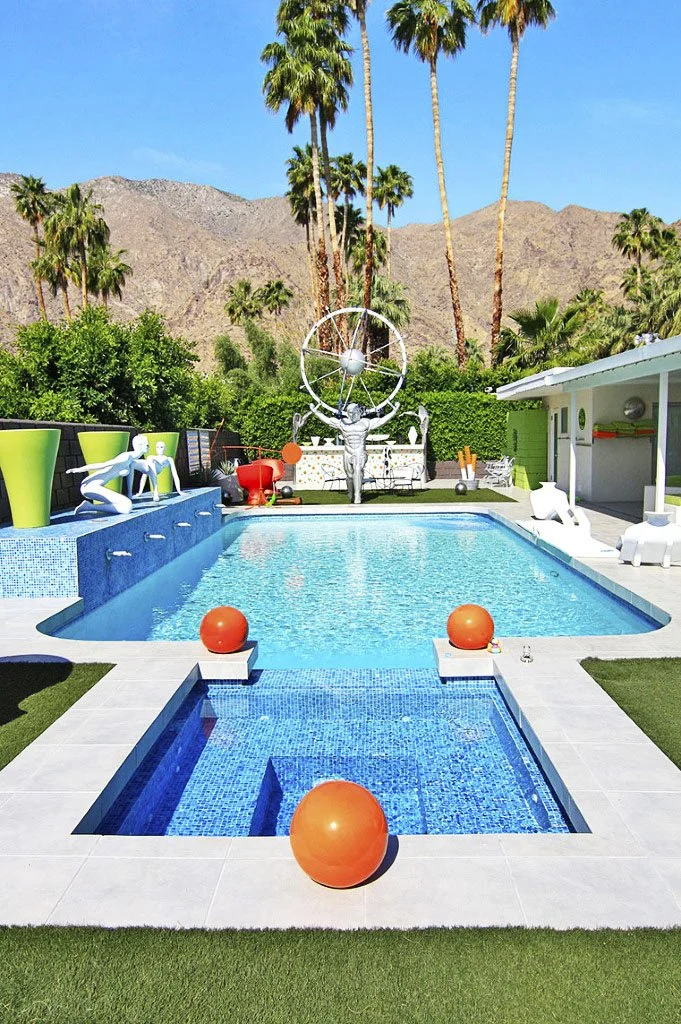 A modern backyard with a swimming pool featuring orange balls floating on water, surrounded by palm trees and a mountain backdrop. Sculptures and patio furniture are visible in the landscaped area.