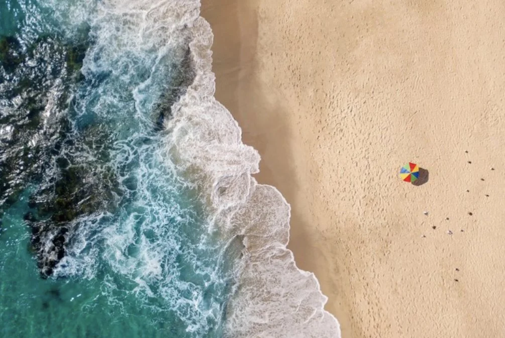 Aerial view of a beach with turquoise waves crashing onto the sand, a colorful umbrella on the right side, and small groups of people scattered around.