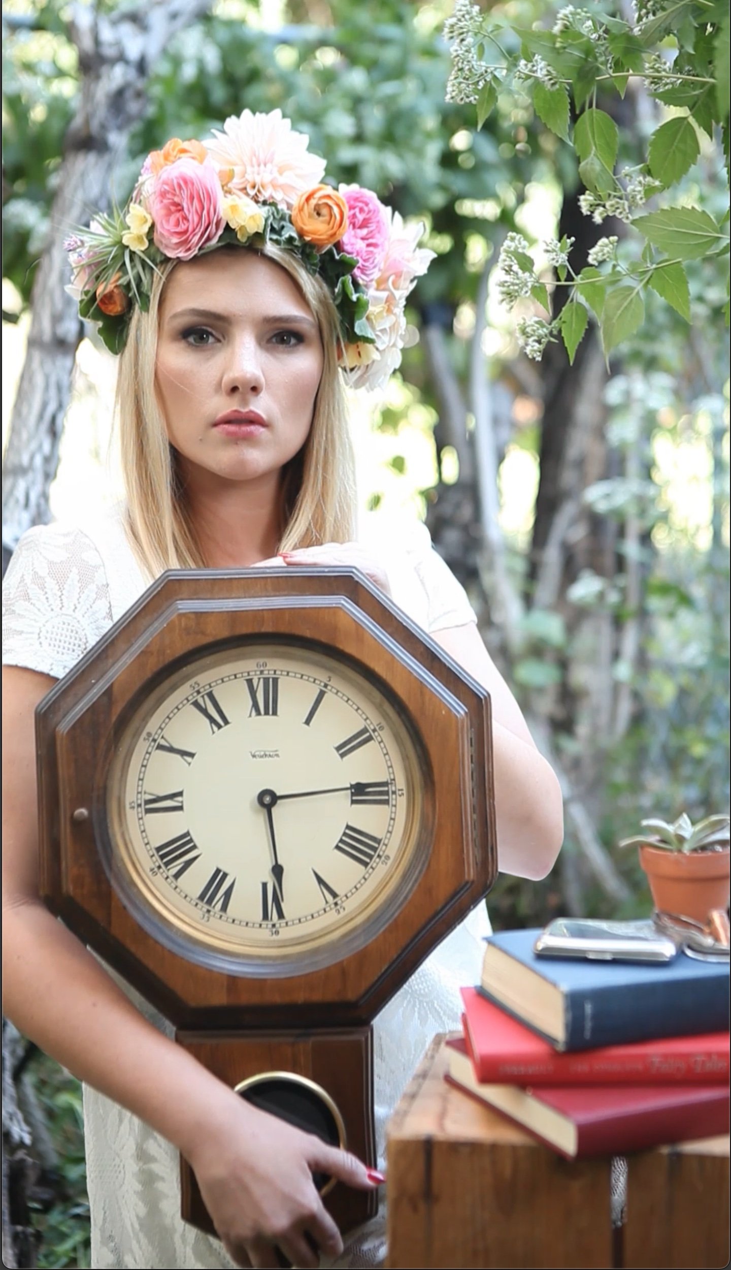 Woman with flower crown holding vintage wall clock in outdoor setting, surrounded by books and a plant.