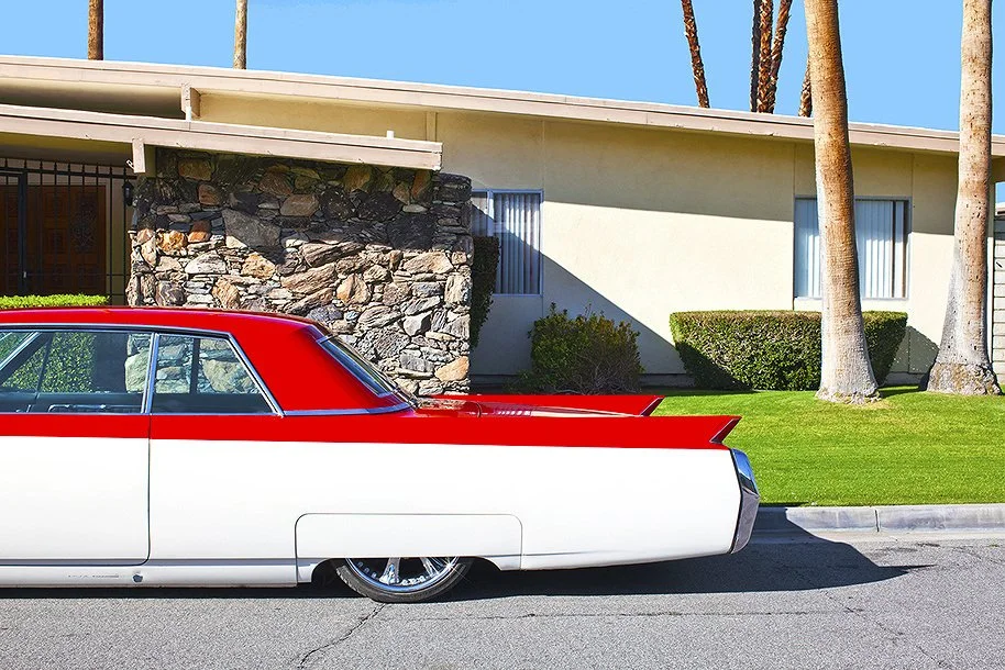 Vintage red and white car parked in front of a mid-century modern house with stone facade and palm trees in the background.