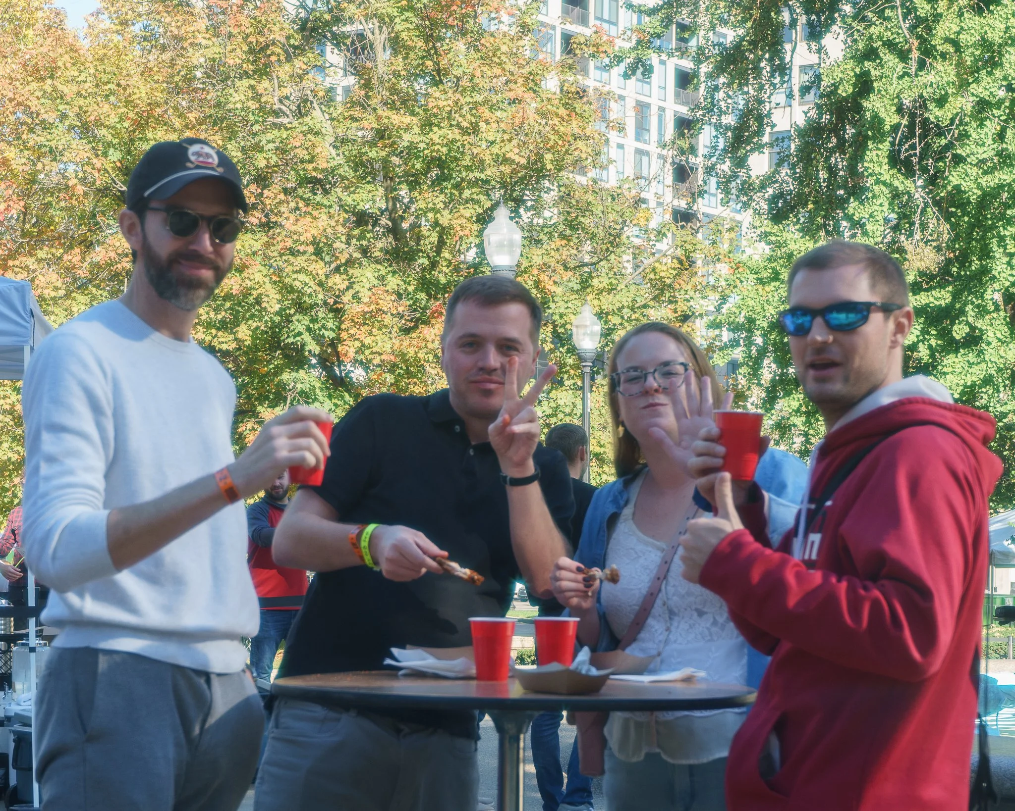 Group of friends at an outdoor gathering, holding red cups and eating snacks. They're smiling and making hand gestures, surrounded by trees and a building in the background.