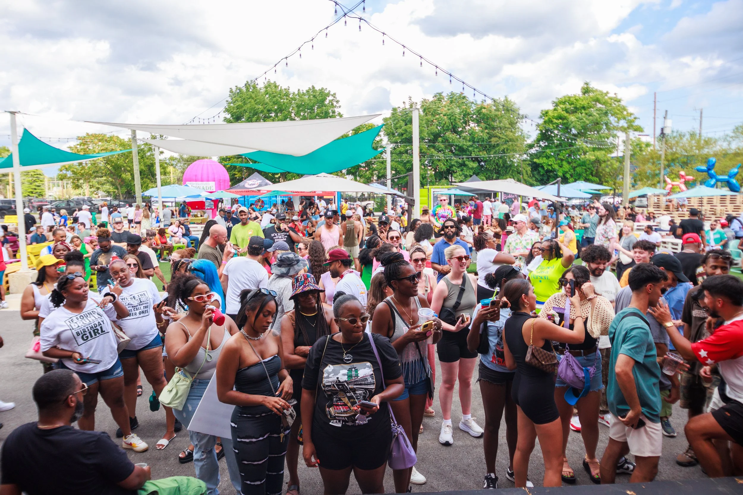 Outdoor festival with a large crowd, food vendors, colorful tents, and people socializing under string lights on a sunny day.