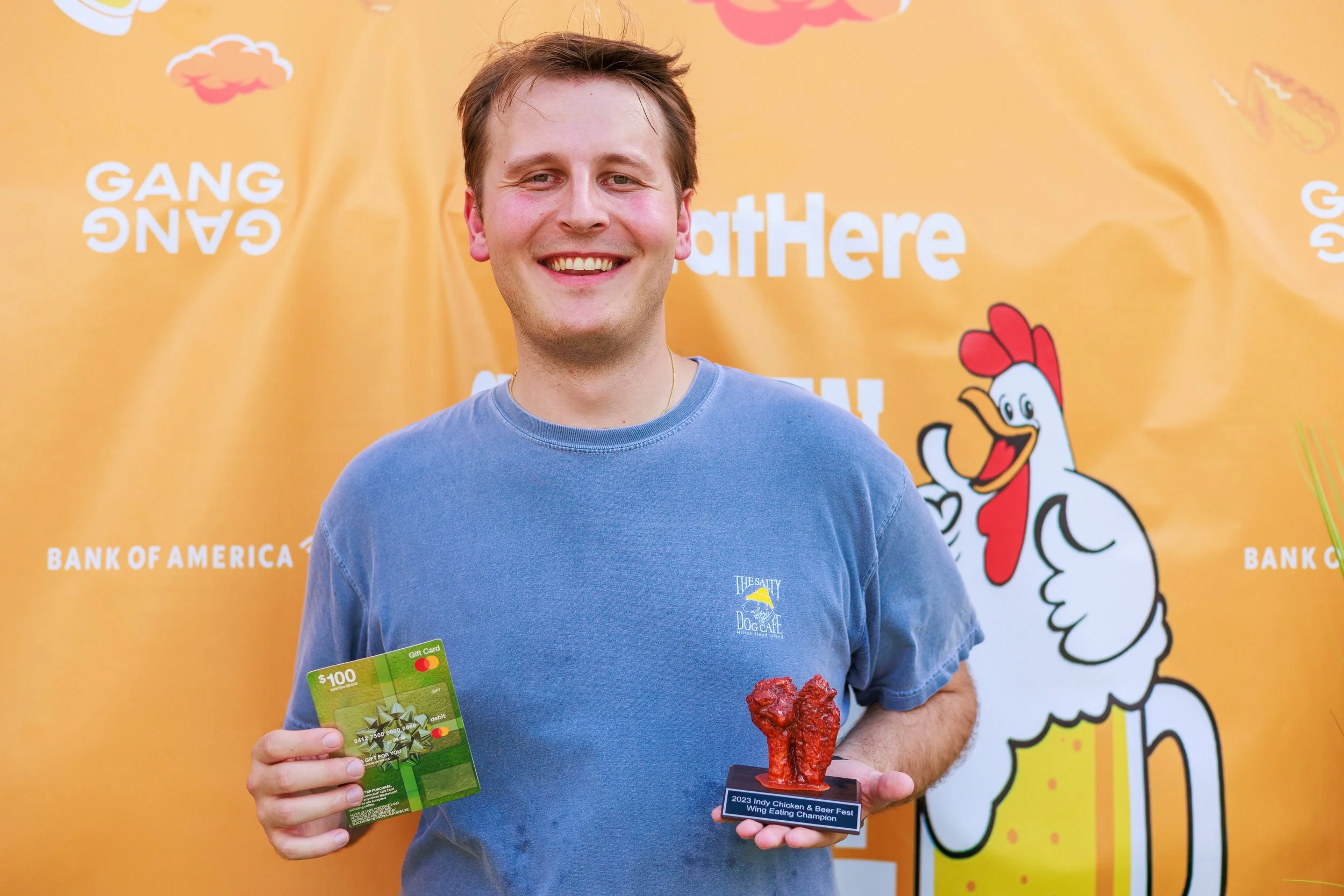 Man holding a $100 gift card and a trophy for winning the 2023 Indy Chicken & Beer Fest wing eating contest, standing in front of an orange backdrop with event logos.