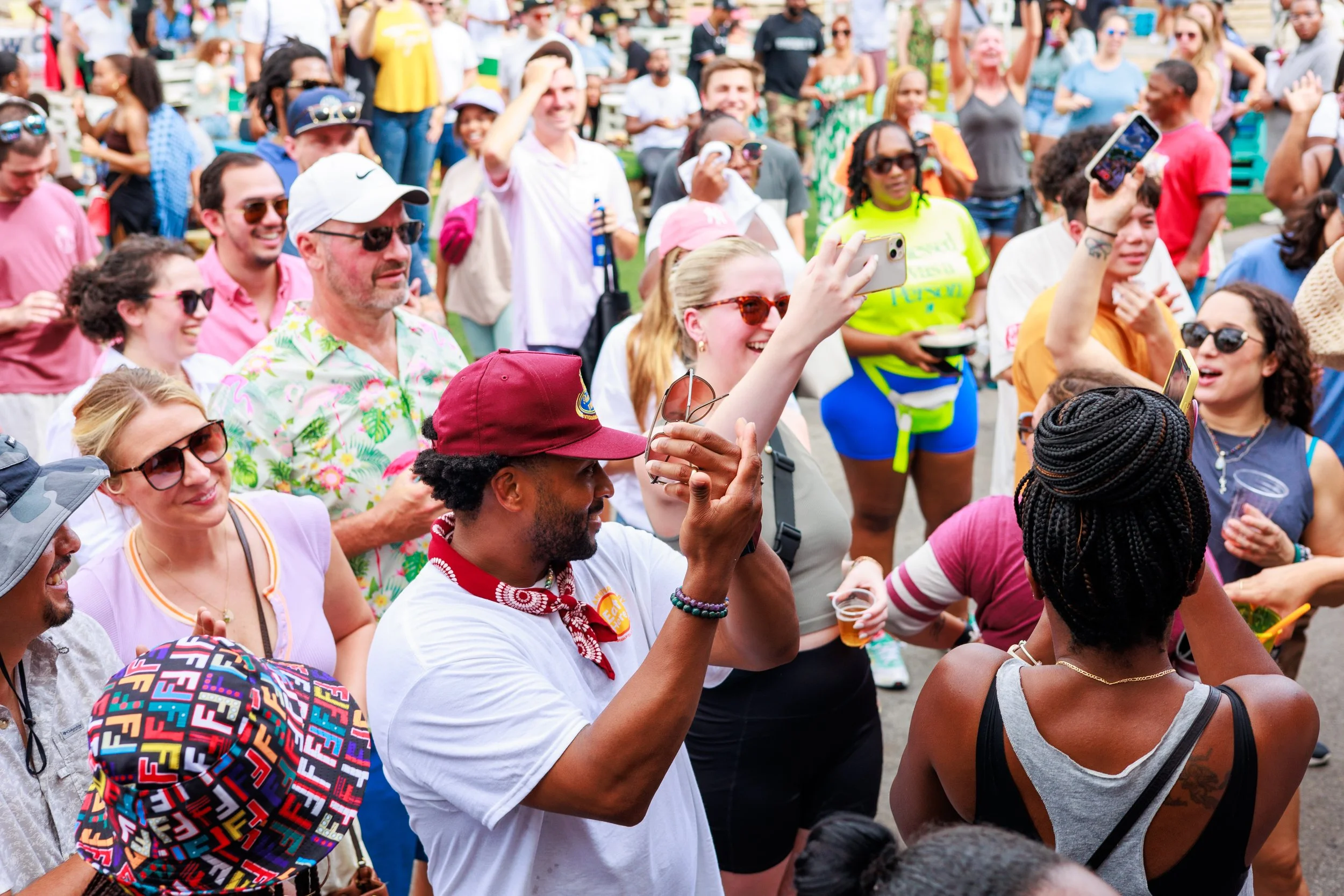 A diverse group of people at an outdoor event, smiling and taking photos with smartphones. Some are wearing sunglasses and hats, while a man in a red cap is in the foreground clapping. The crowd is lively and appears to be enjoying the occasion.