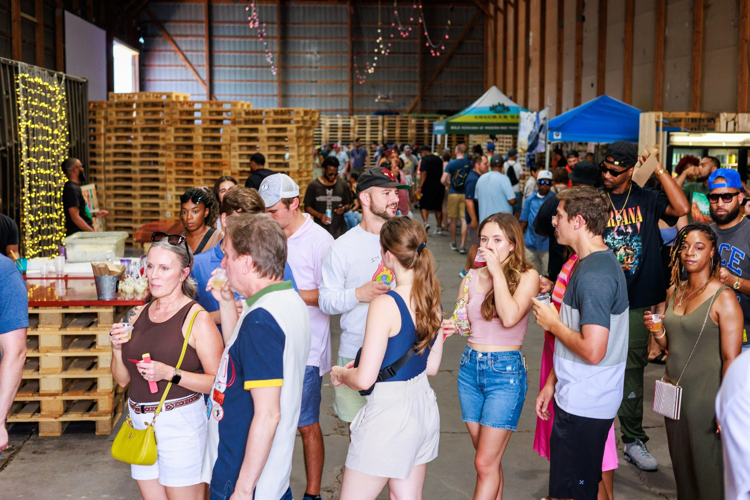 People at a warehouse event holding drinks, chatting, and gathered around wooden pallets and booths.