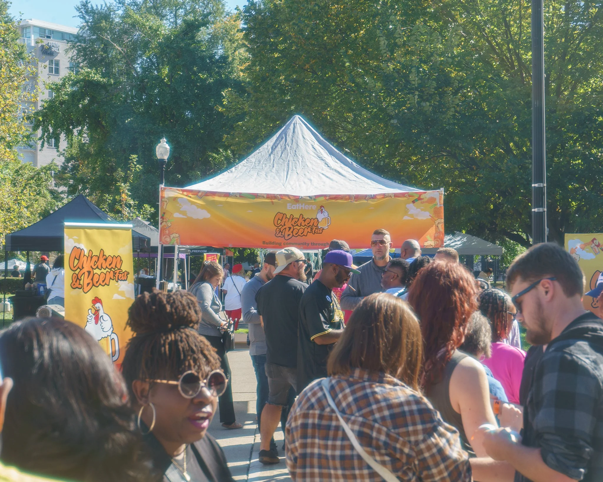 People gathered at outdoor event with tents, "Chicken & Beer Fest" signs, and trees in background.