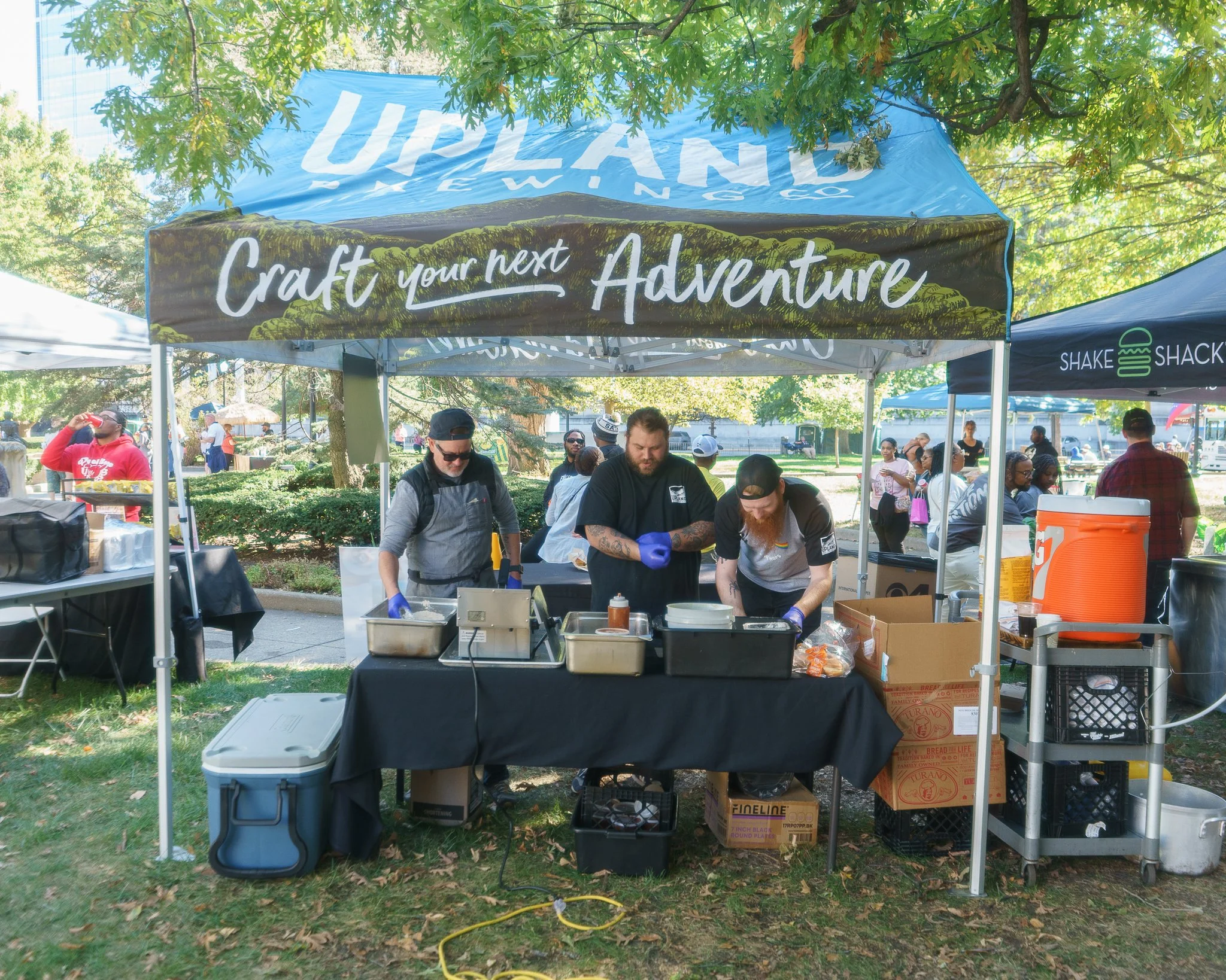 Outdoor food booth with three people preparing food under a tent labeled "Upland Brewing Co." at a festival. The booth has cooking equipment, boxes, and a cooler. Nearby, a "Shake Shack" setup is visible with a bright orange cooler. People are in the background enjoying the event under trees.