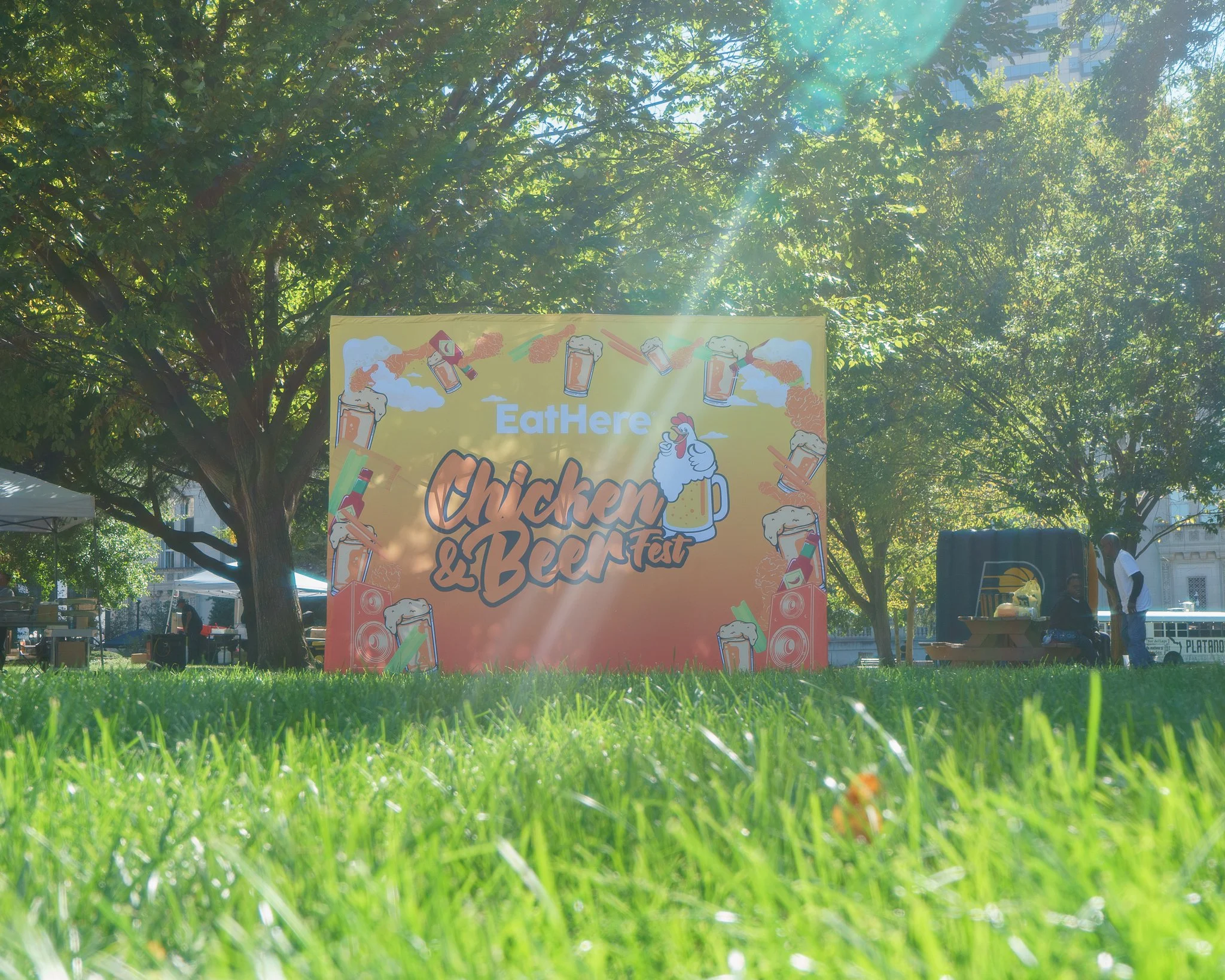 Outdoor sign in a park for the "Chicken & Beer Fest" by EatHere, featuring illustrations of chicken and beer items, with trees and picnic setup in the background.