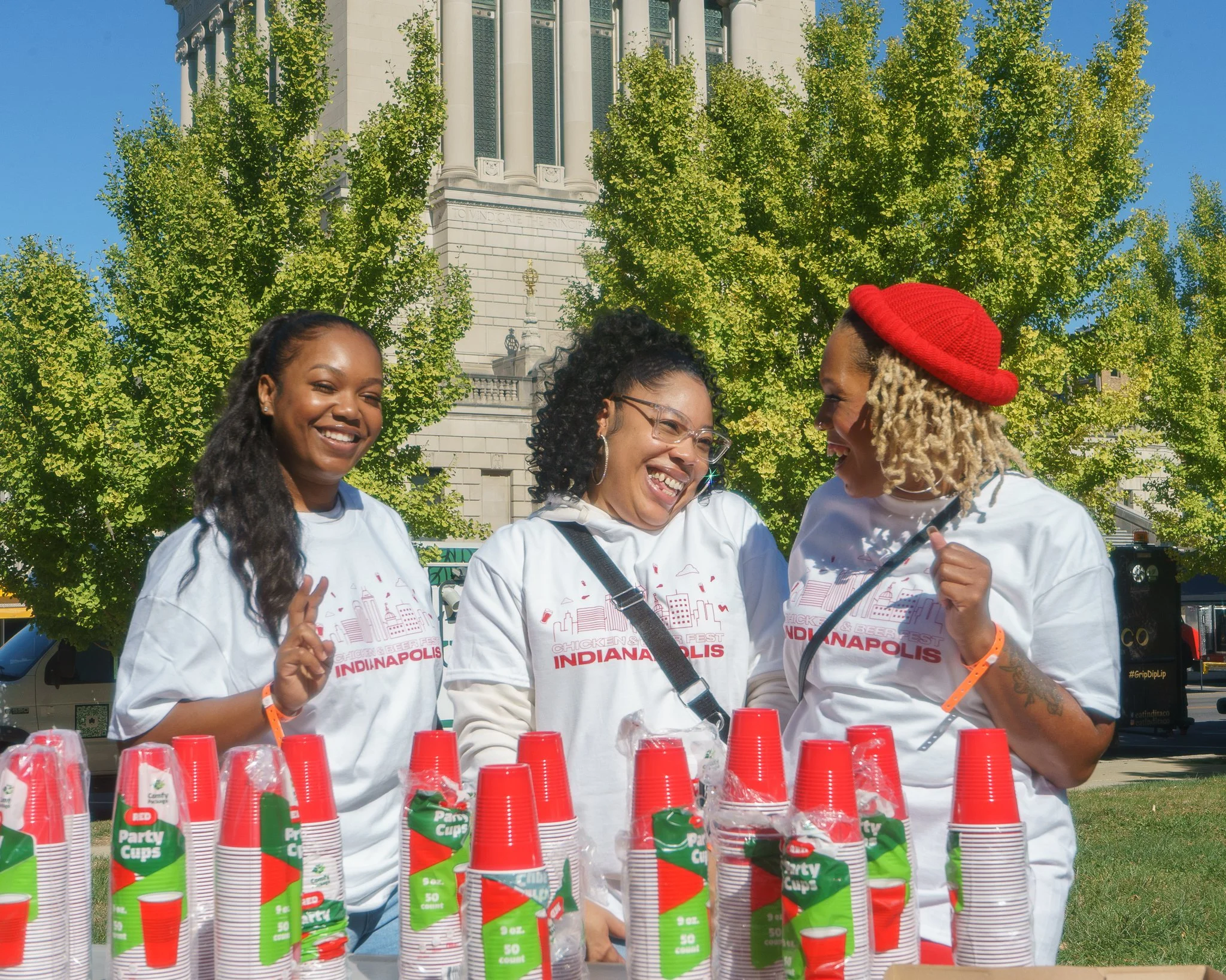 Three women in matching Indianapolis t-shirts posing with red party cups at an outdoor event.