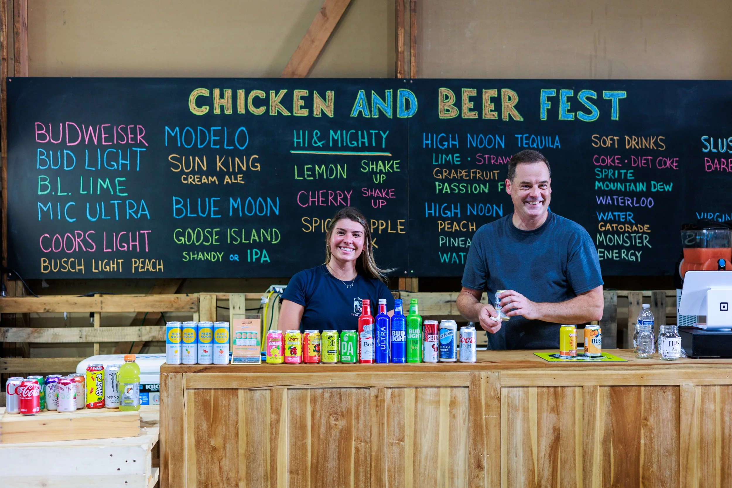 Two people standing behind a wooden bar counter at a 'Chicken and Beer Fest,' with a variety of beer cans and bottles displayed. A chalkboard behind them lists drink options, including various beers, soft drinks, and cocktails.